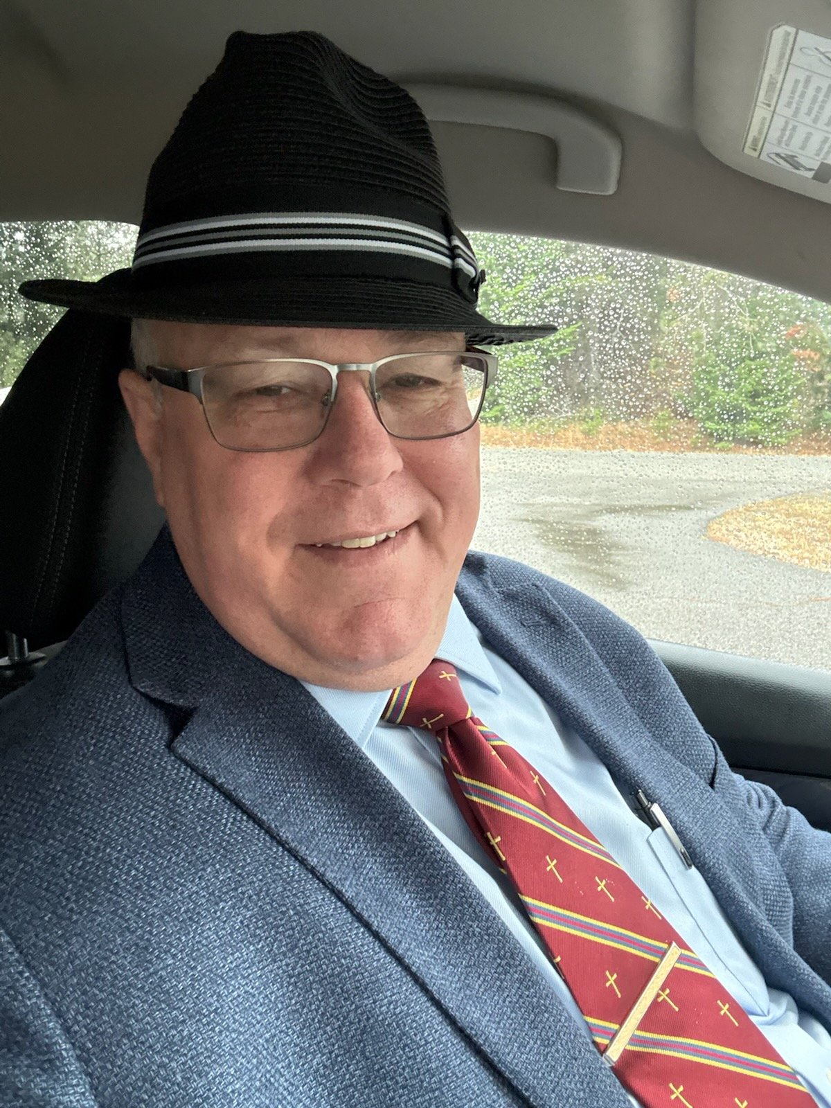 A man wearing glasses, a blue suit, a red tie with yellow crosses, and a black fedora hat with white stripes, sitting inside a car with raindrops on the window.