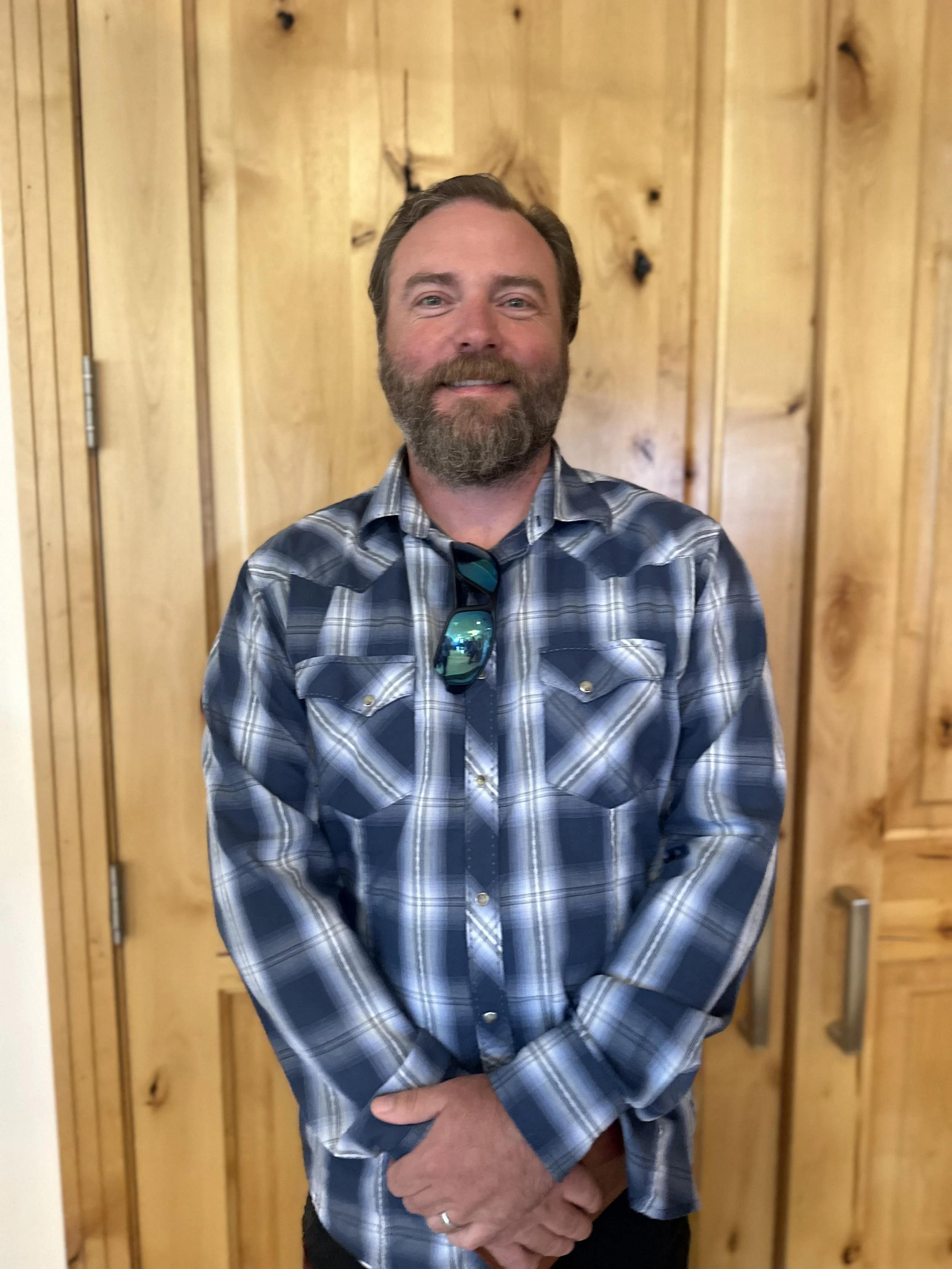 A middle-aged man with a beard, wearing a blue plaid shirt, smiling, and standing in front of wooden paneling. He has sunglasses hanging from his shirt collar.
