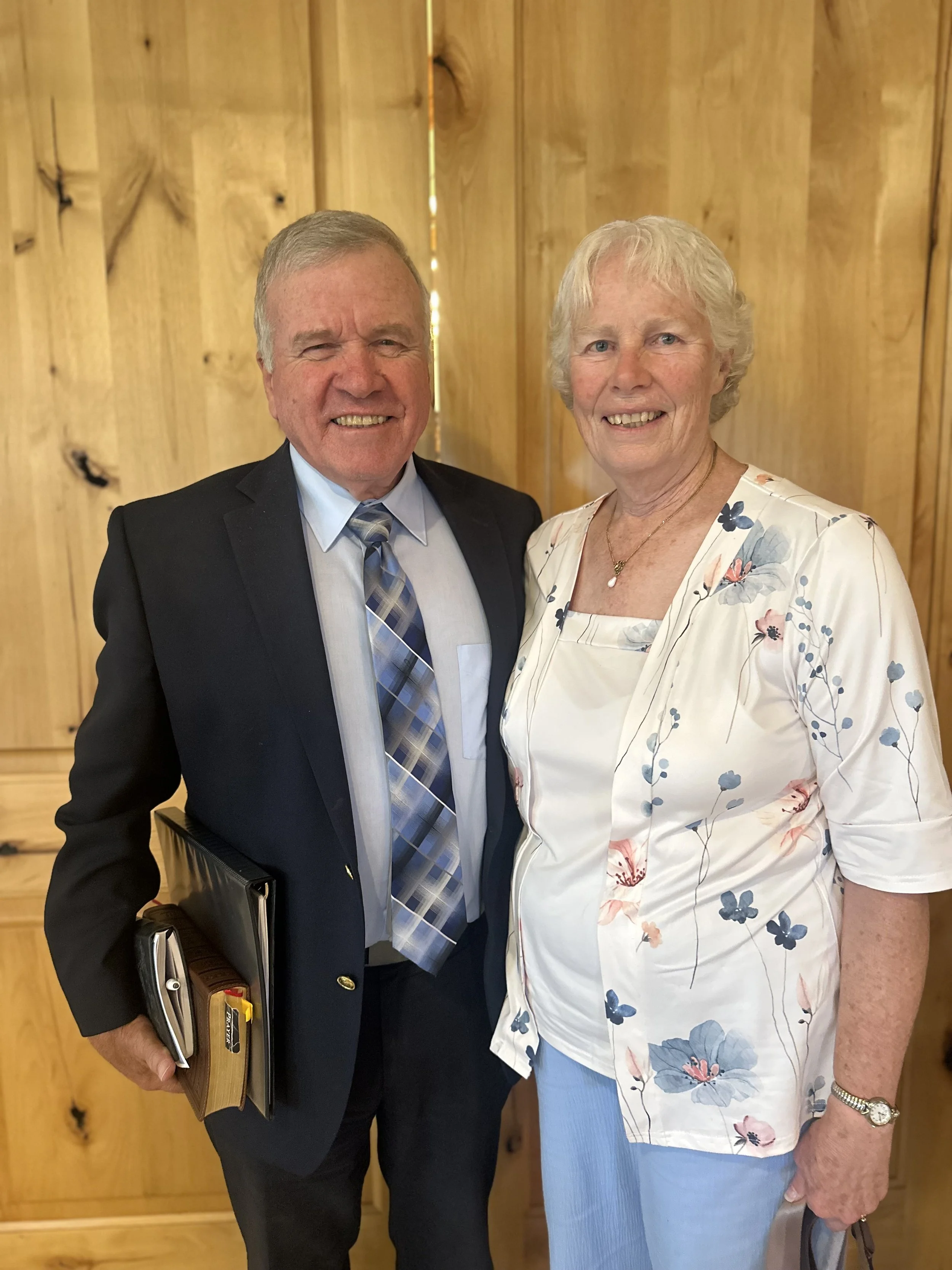 A smiling older woman with short blonde hair wearing a pink jacket and floral blouse, standing next to a smiling middle-aged man with short gray hair wearing a beige suit and striped tie, in front of wooden paneling.