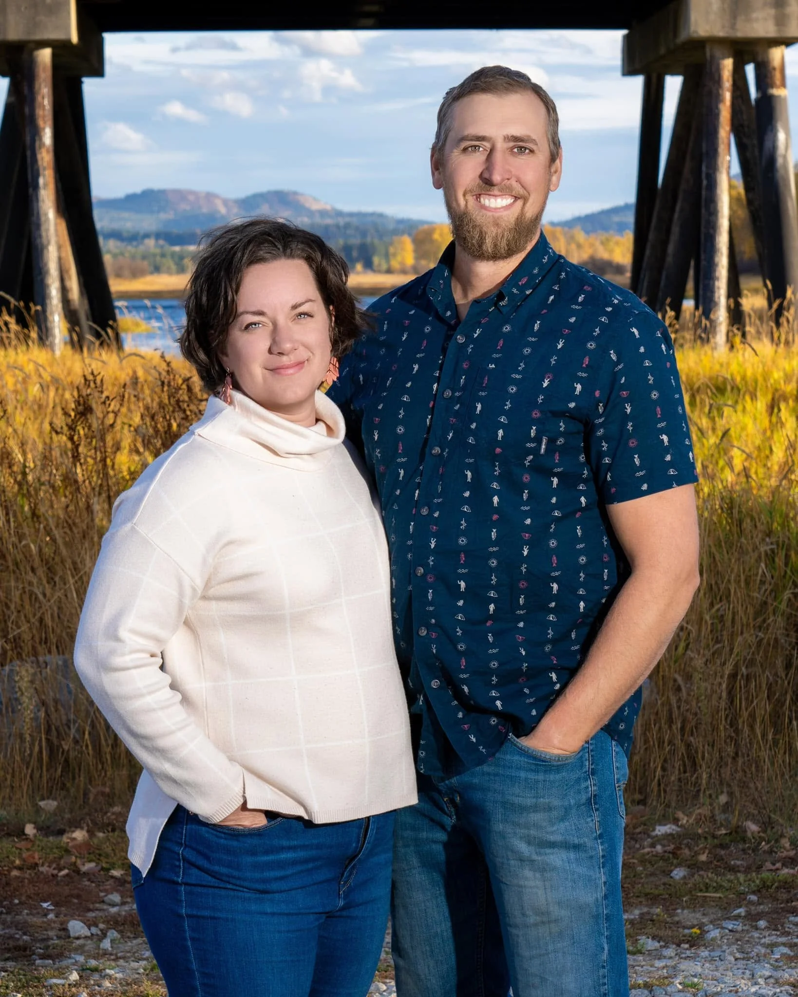 A smiling man and woman stand close together outdoors near a wooden bridge, with mountains, blue sky, and clouds in the background during fall.