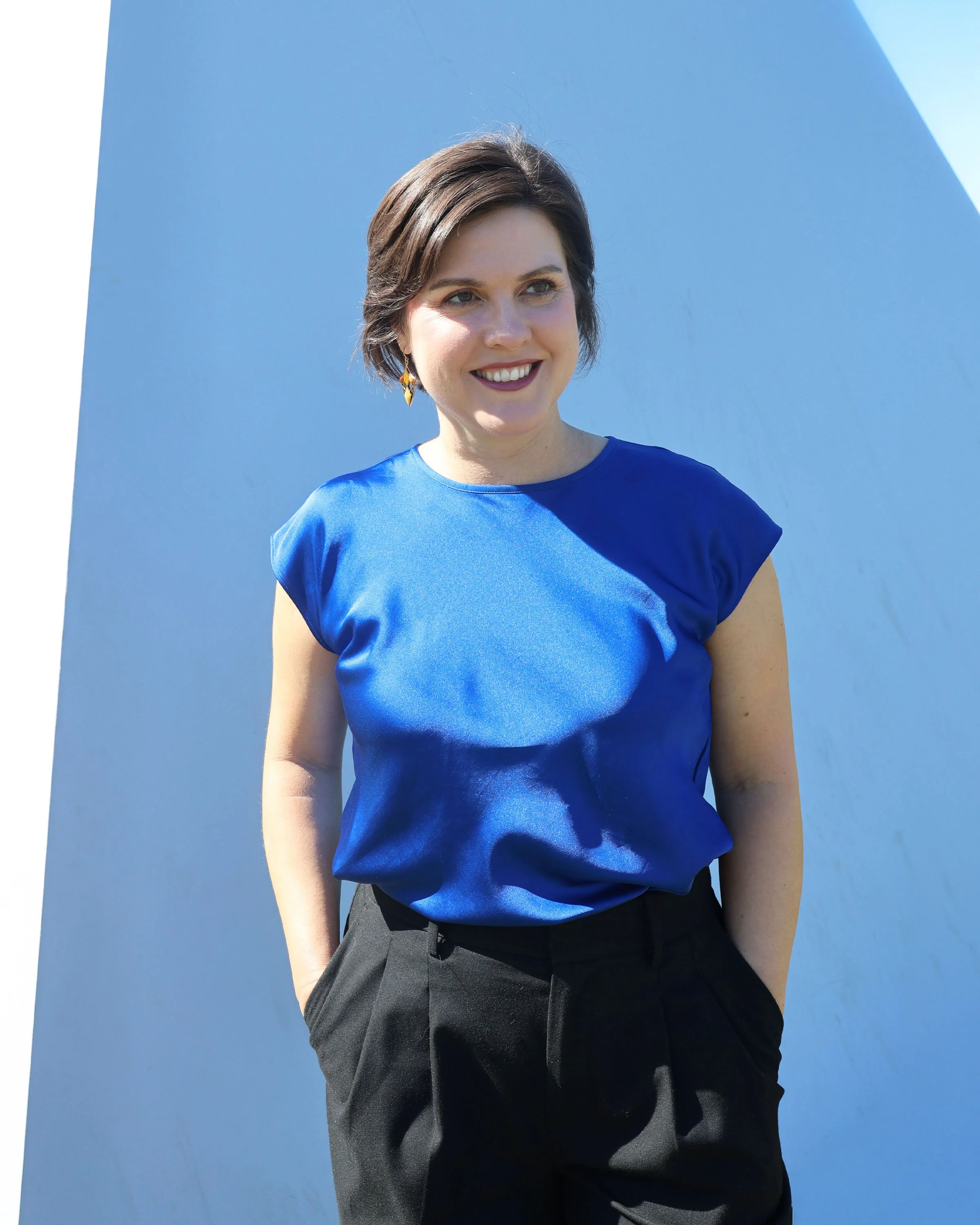 Young woman with short brown hair, wearing a blue top and black pants, standing outdoors against a bright blue sky with a white, curved wall in the background, smiling and looking to her right.