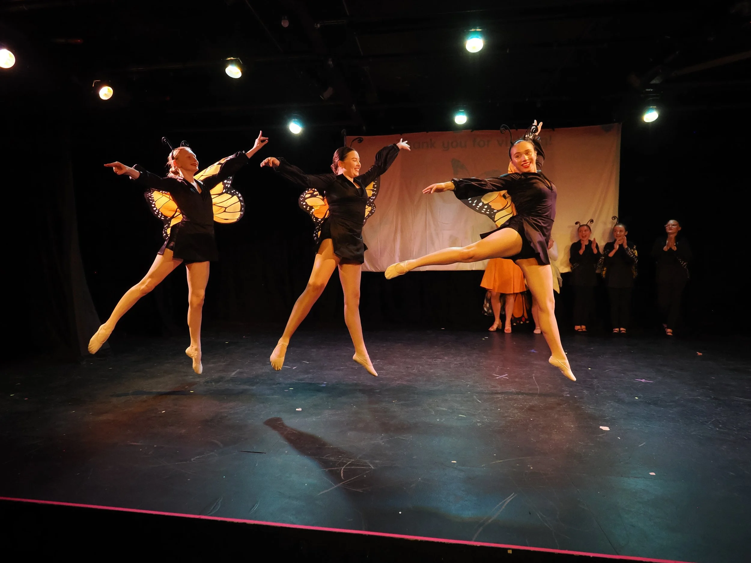Three female dancers dressed as butterflies in black costumes with butterfly wings, performing on stage, with a woman in the background applauding.