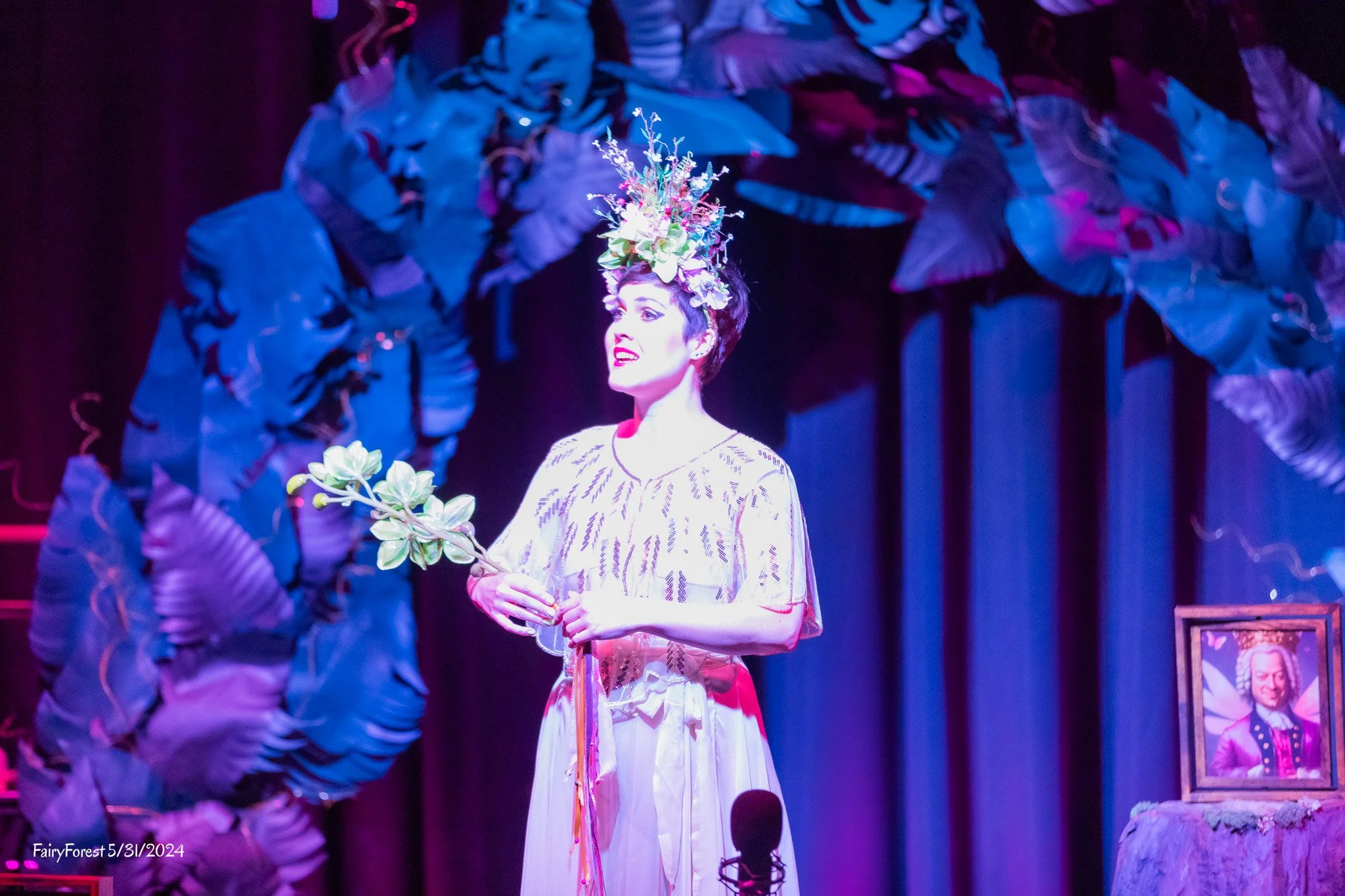 A woman wearing a light-colored dress with black patterns and a large floral headpiece made of green leaves and pink, purple, and white flowers stands on stage holding a small bouquet. The stage background features large blue and purple leaves, and there are framed photographs on the stage leg. The lighting is purple and pink, creating a vibrant atmosphere. Text at the bottom left corner reads 'FairyForest 5/31/2024'.