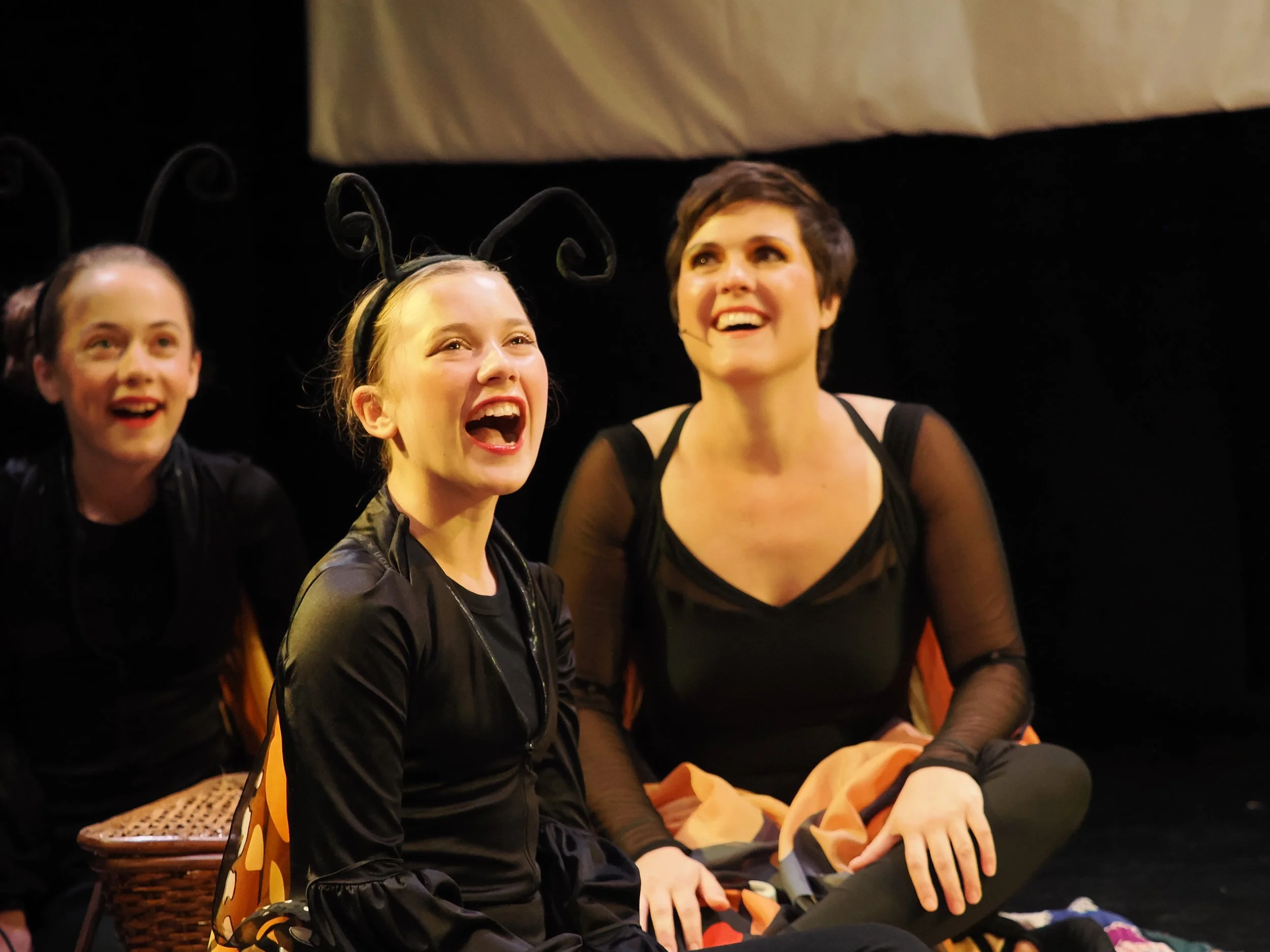 Three women seated on stage, smiling and enjoying a performance. The woman in the center is wearing bee antennae headband, the woman on the right has short dark hair, and the woman on the left has dark hair tied back. The woman in the center is wearing a black jacket, and the woman on the right is wearing a sheer black top. They are sitting on chairs with a dark background above them.