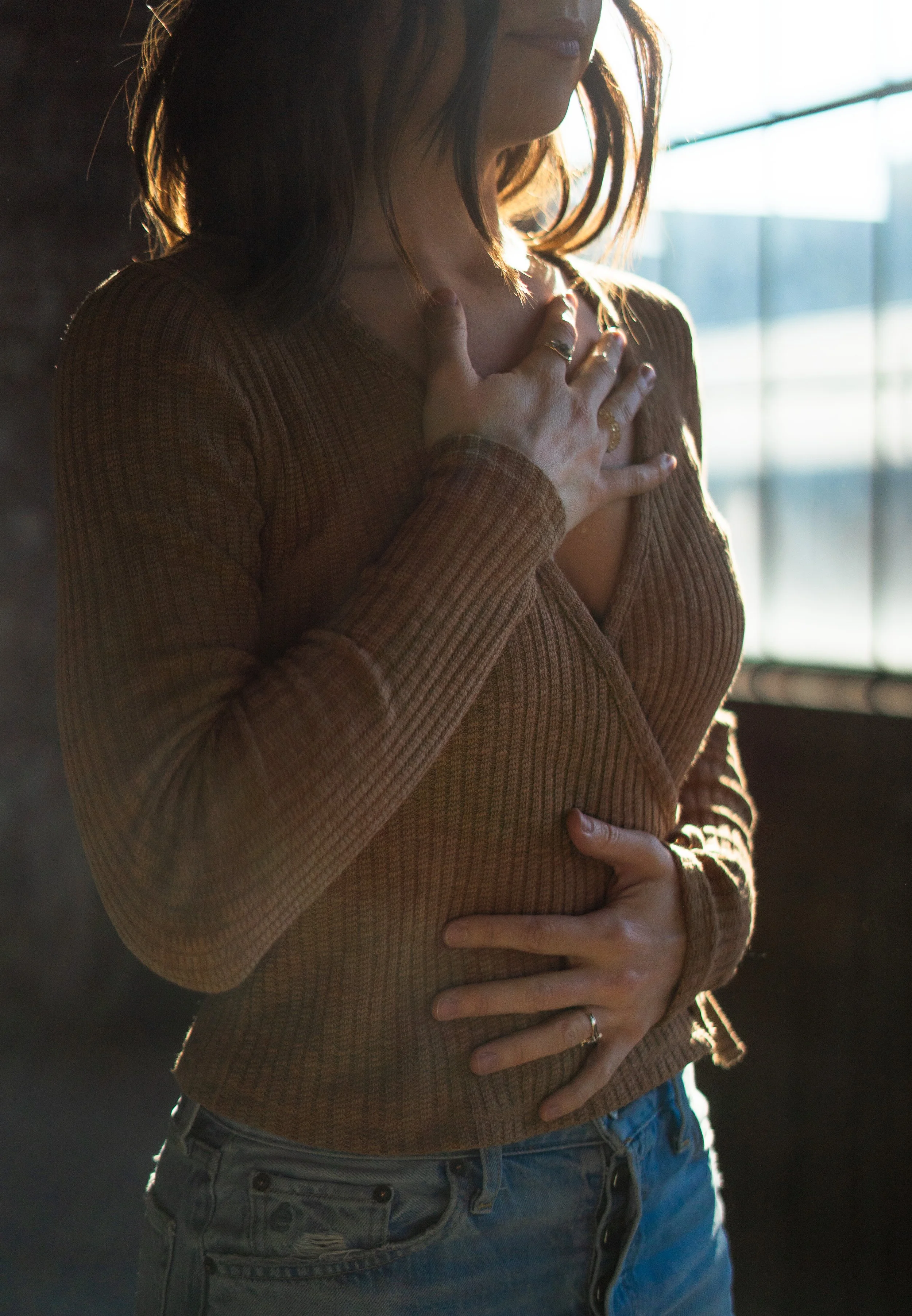 A woman with brown hair wearing a brown long-sleeve shirt and blue jeans, with hands on the heart space and stomach, standing near a window with sunlight streaming in.