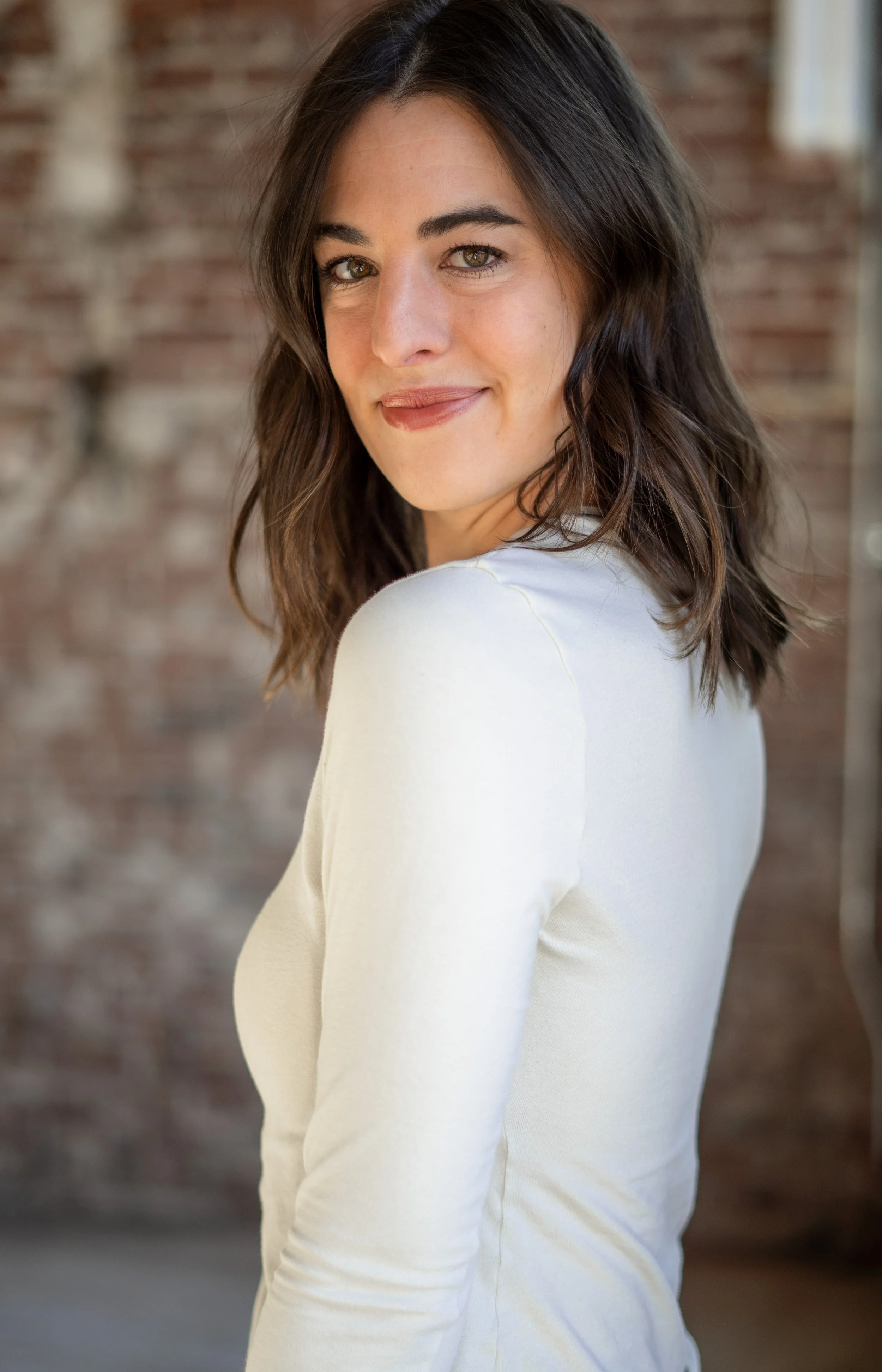 A woman with shoulder-length dark brown hair and hazel eyes, smiling softly, wearing a white long-sleeve shirt, standing in front of a blurred brick wall background.