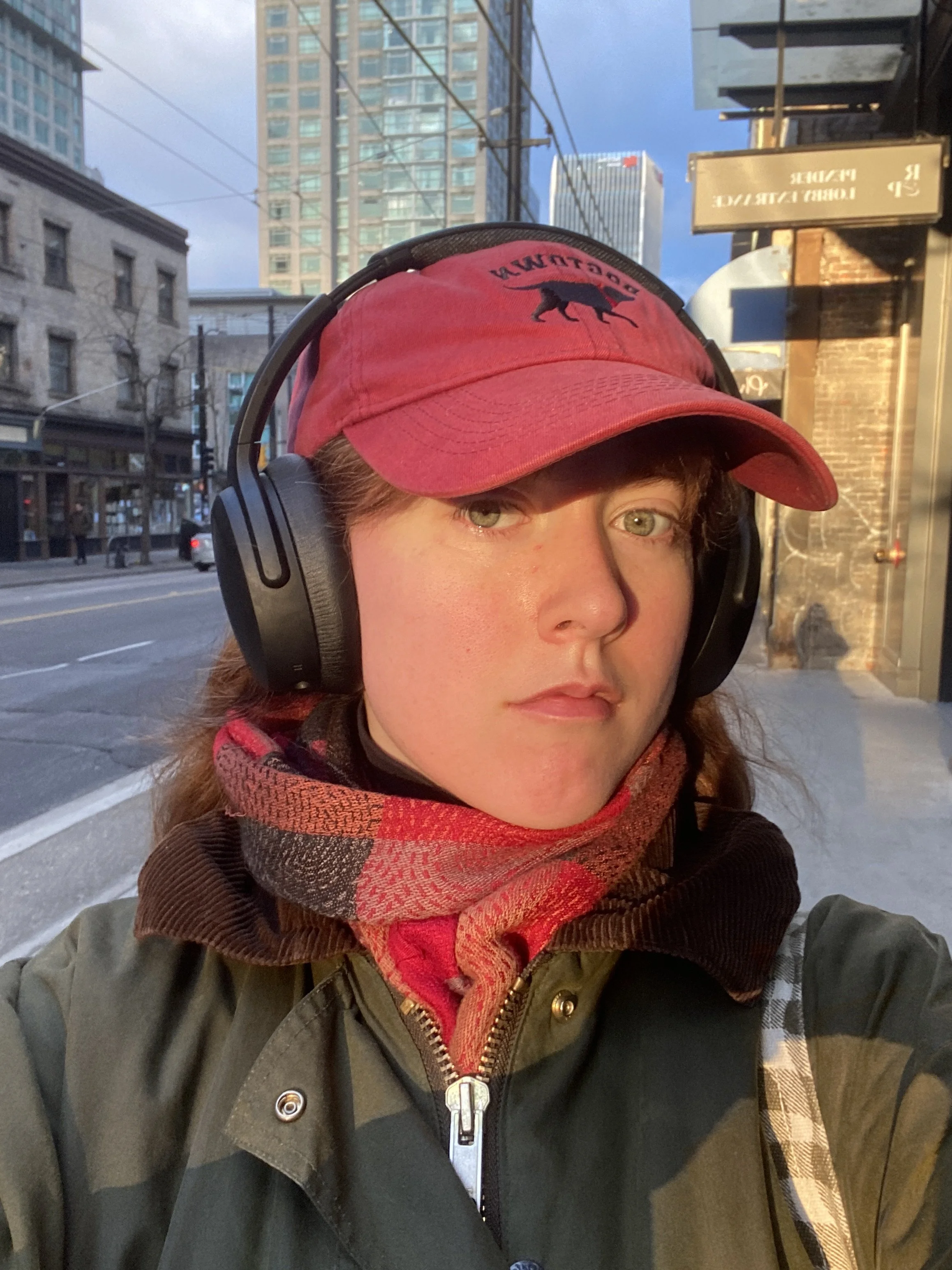 Close-up selfie of a person wearing a red baseball cap, headphones, and a multicolored scarf, standing outdoors in an urban setting during daylight with high-rise buildings and city street in the background.