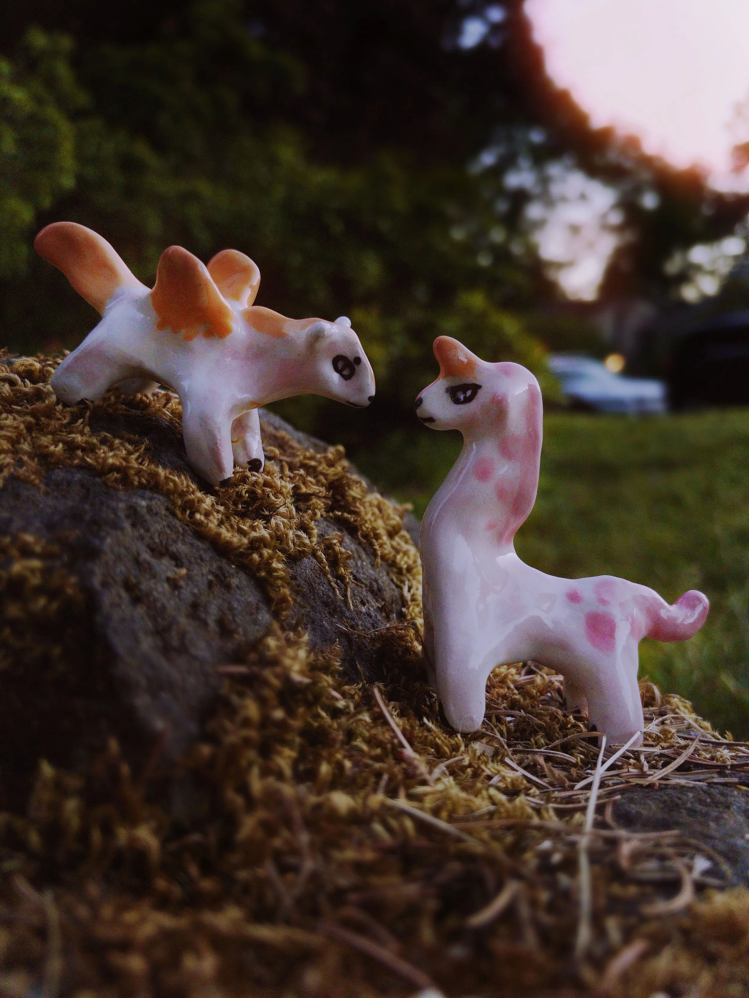 Two small, ceramic figurines with pink and orange accents on a mossy surface outdoors during sunset. One is a unicorn with a long neck, the other on the left standing higher on the rock is a pegasus.