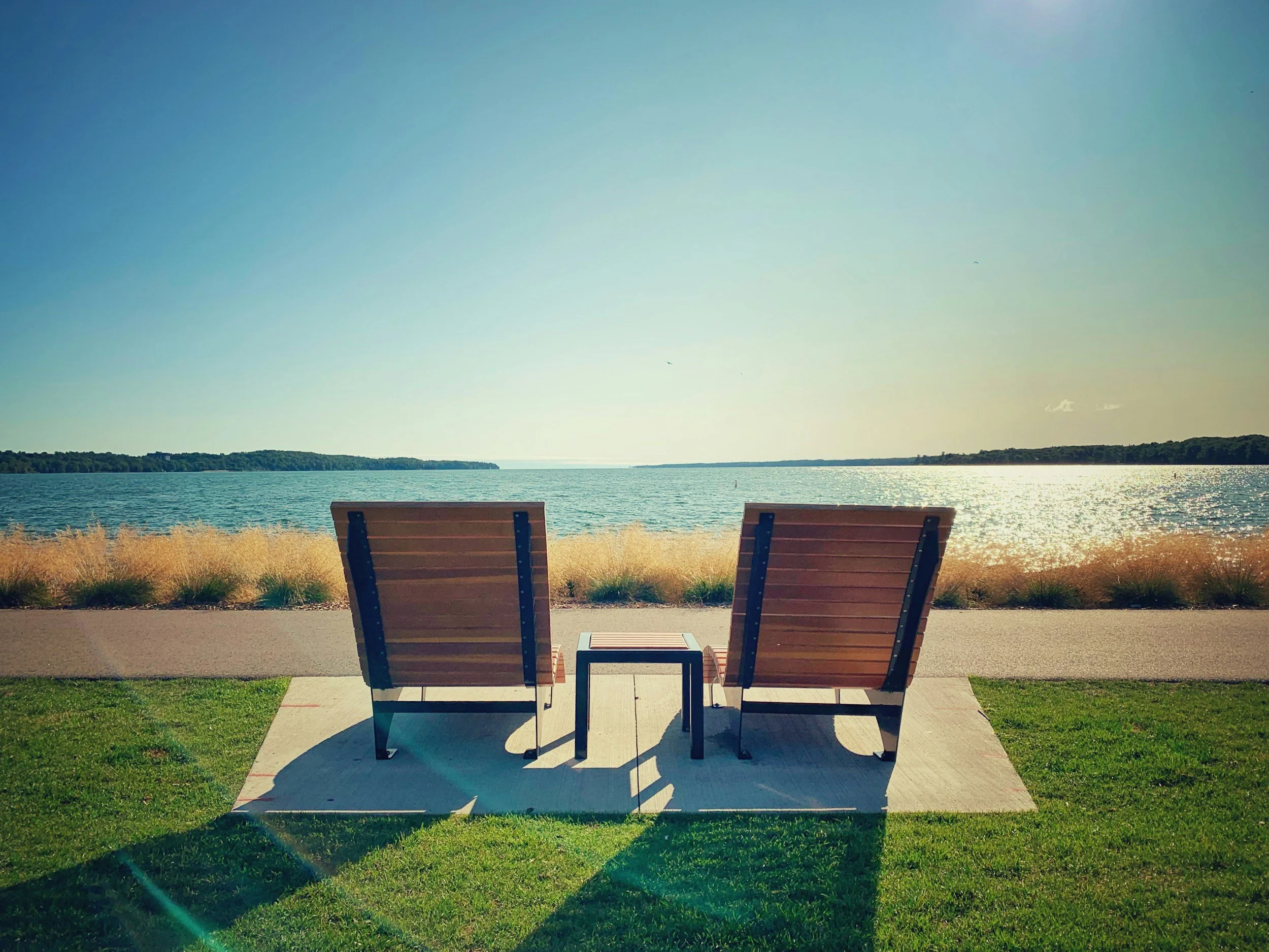 Two empty wooden benches facing Lake Simcoe with a tree-lined shoreline, under a clear sky.