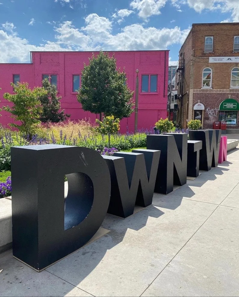 Large black 3D letters spelling 'DOWNTOWN' on a sidewalk, with a pink building, trees, and a blue sky with clouds in the background.