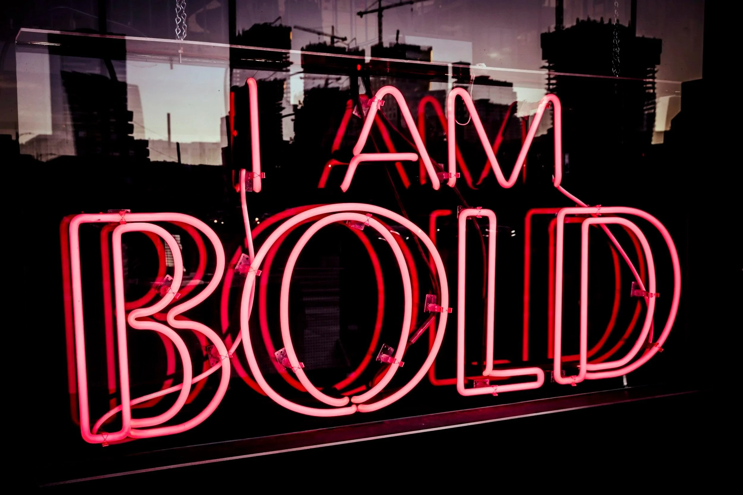 Neon sign with red letters spelling 'I AM BOLD' reflected in a glass window.