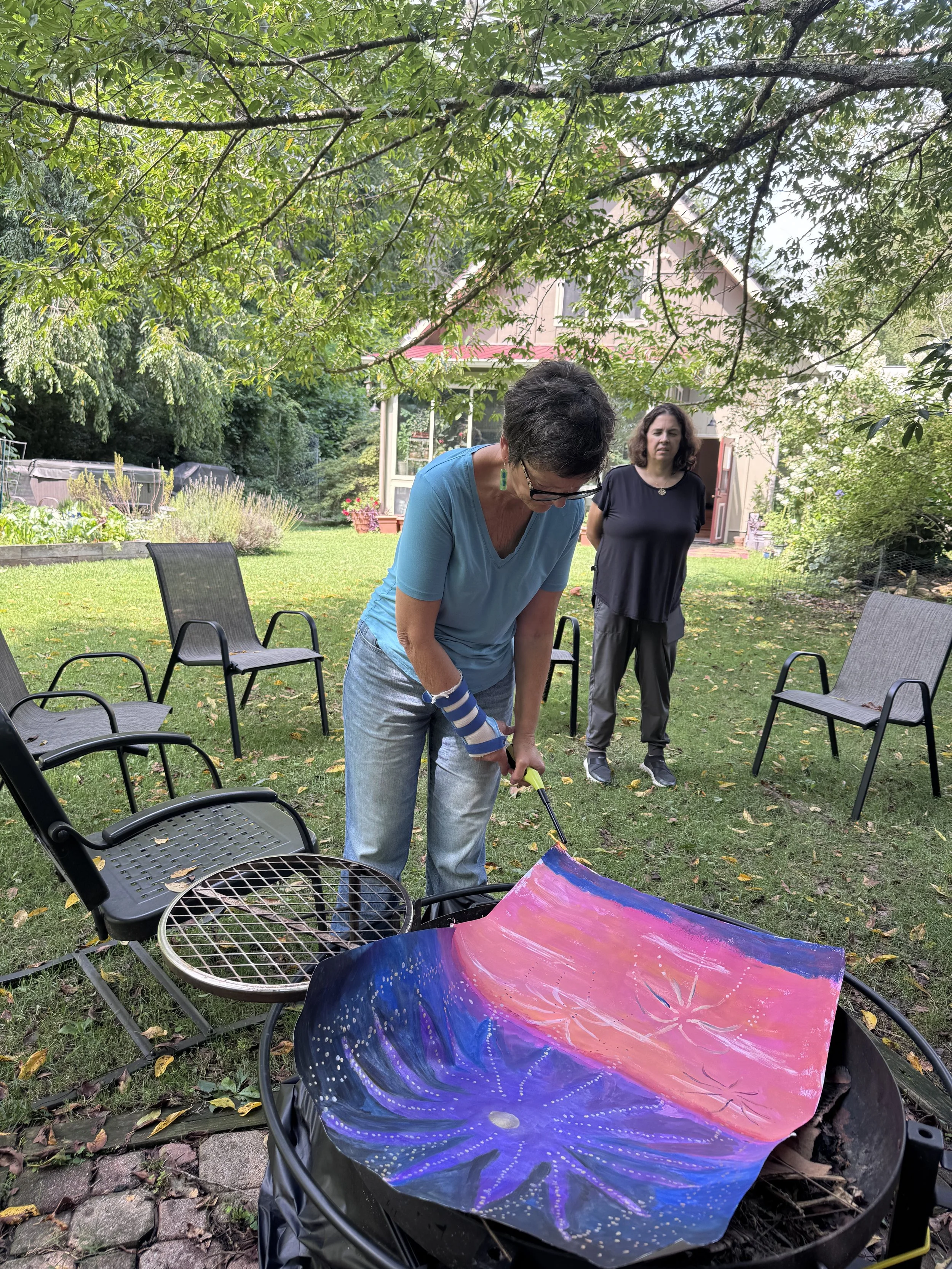 A woman in a blue shirt is painting a large colorful galaxy-themed artwork on a circular surface outdoors, with another woman observing in the background. There are chairs surrounding a garden area with trees and plants.