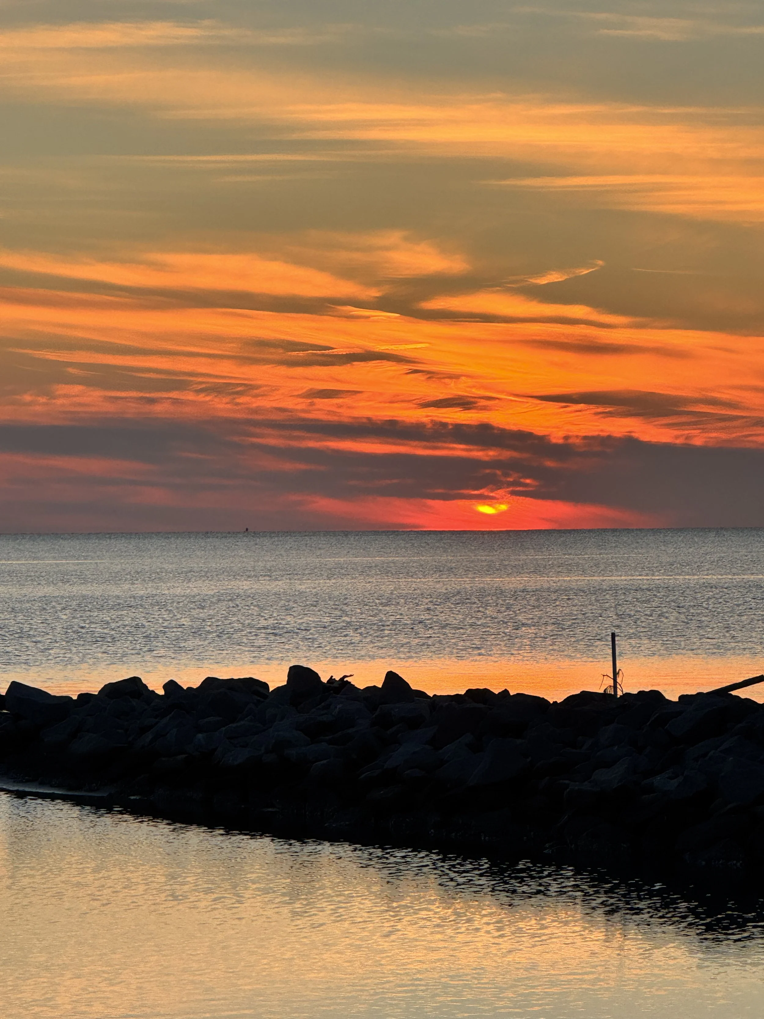 A peaceful sunset over a calm body of water with a rocky shoreline in the foreground.