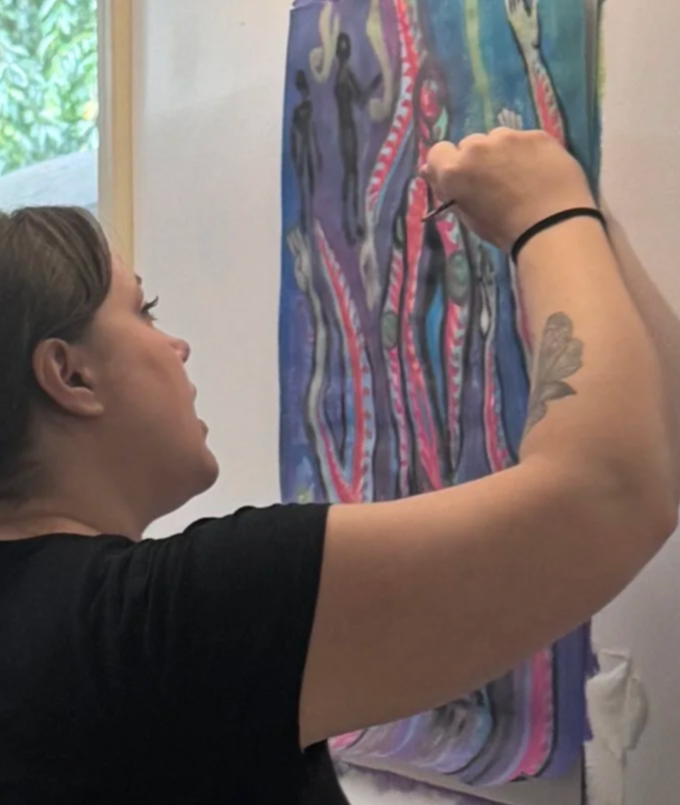 A woman with brown hair, wearing a black shirt, is painting a colorful abstract piece on a wall.