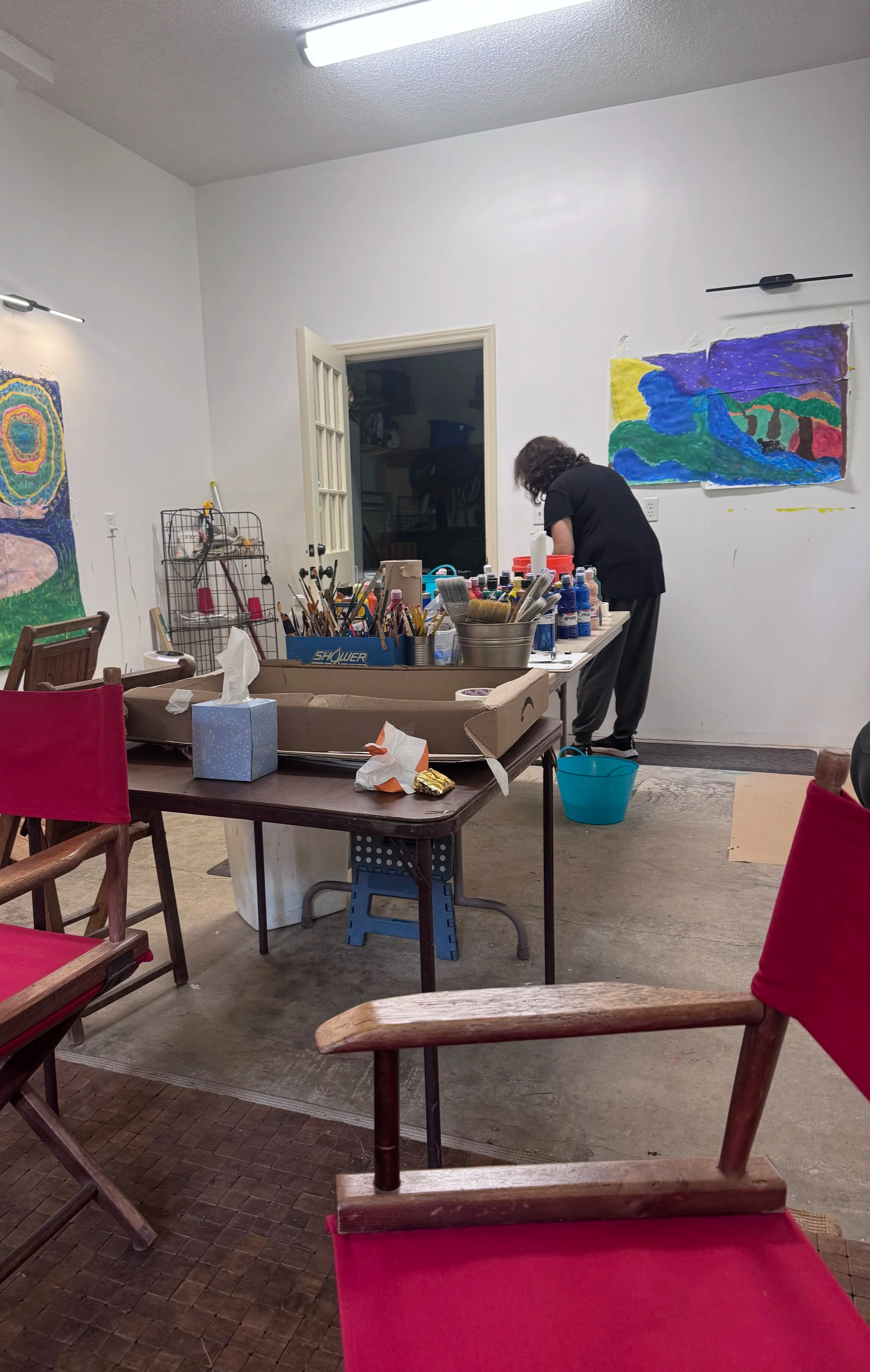 A person with curly hair, wearing a black shirt and gray pants, is working at a table covered with art supplies in a room with white walls. The room has colorful paintings on the wall, a door leading to another room, and red chairs around the table. There are various art tools, paint bottles, and a tissue box on the table.