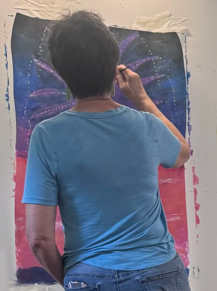An artist painting a vibrant, colorful mural featuring a background with star-like details and purple curved shapes, seen from behind.