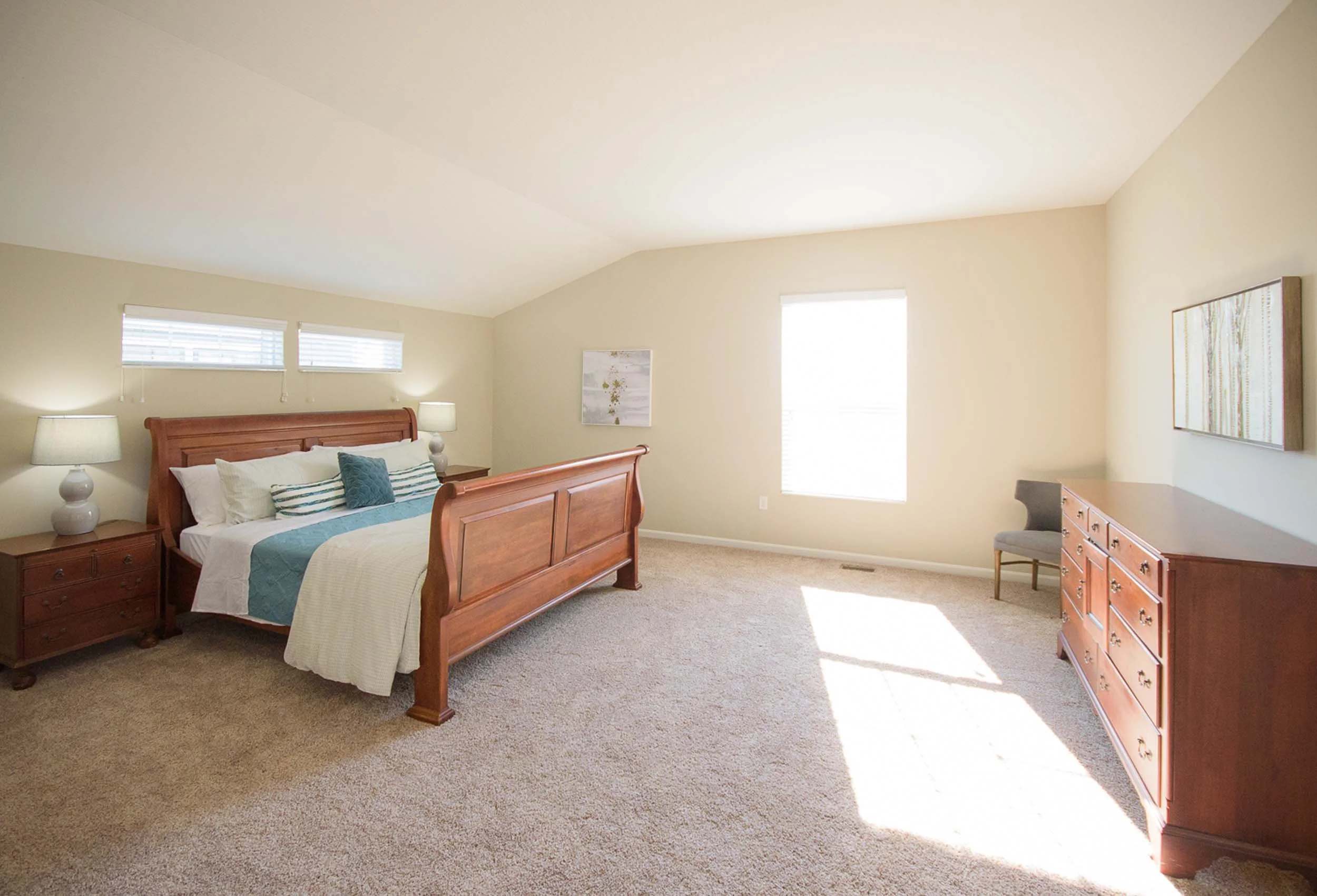 A bedroom with a wooden bed frame, white bedding, and decorative pillows. Two bedside tables with lamps, artwork on the walls, a dresser, and a chair near a window with blinds. Carpeted floor and ample natural light.