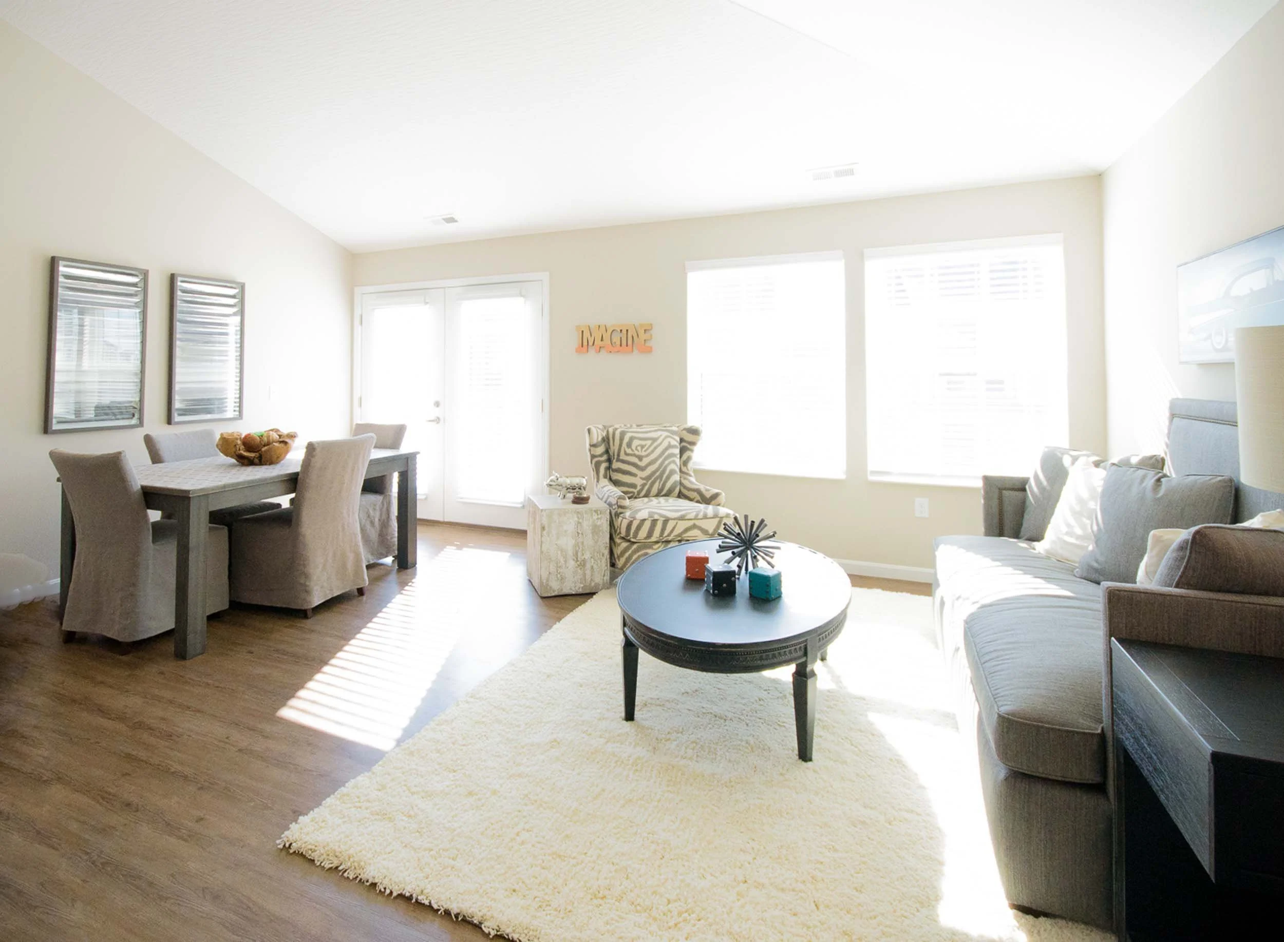 Bright living room with large windows, white walls, beige and gray furniture, a round black coffee table with dice and decorative object, a striped armchair, a beige shaggy rug, and wall art including a wooden "IMAGINE" sign.