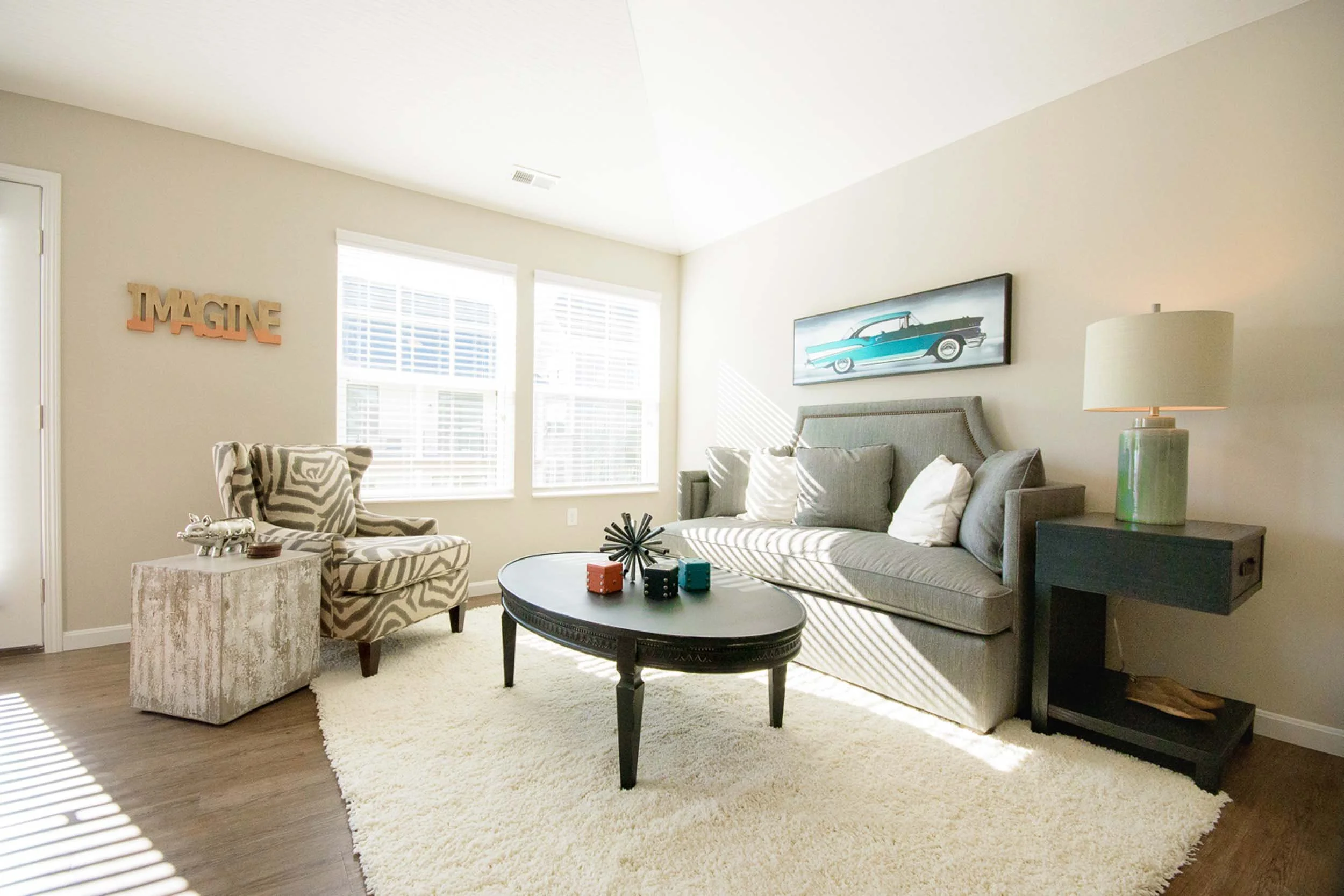 Living room with large windows, a beige wall with decorative words, a patterned armchair, a gray sofa with pillows, a round black coffee table, a table lamp, and wall art of a vintage car.