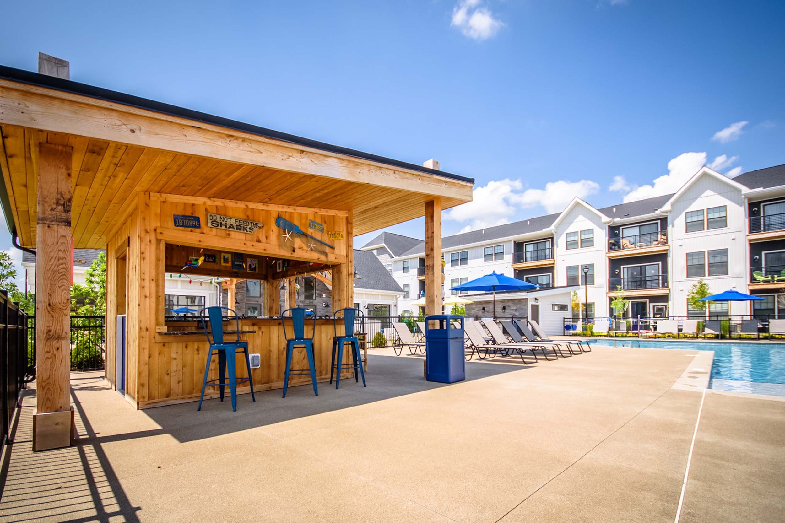A swimming pool area with lounge chairs, a poolside bar with blue barstools, umbrellas, and a multi-story apartment complex in the background on a sunny day.