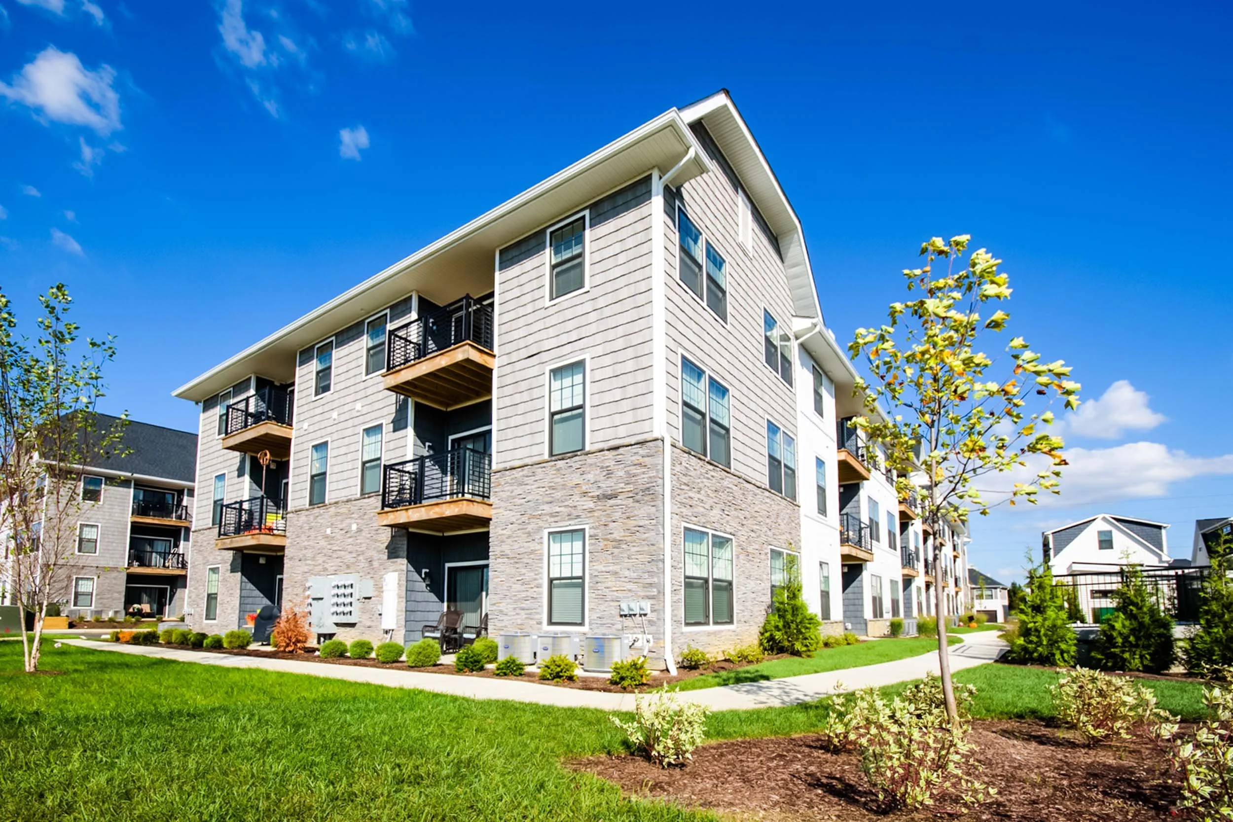 A modern multi-story apartment building with balconies, surrounded by green lawns and young trees, under a bright blue sky with some clouds.
