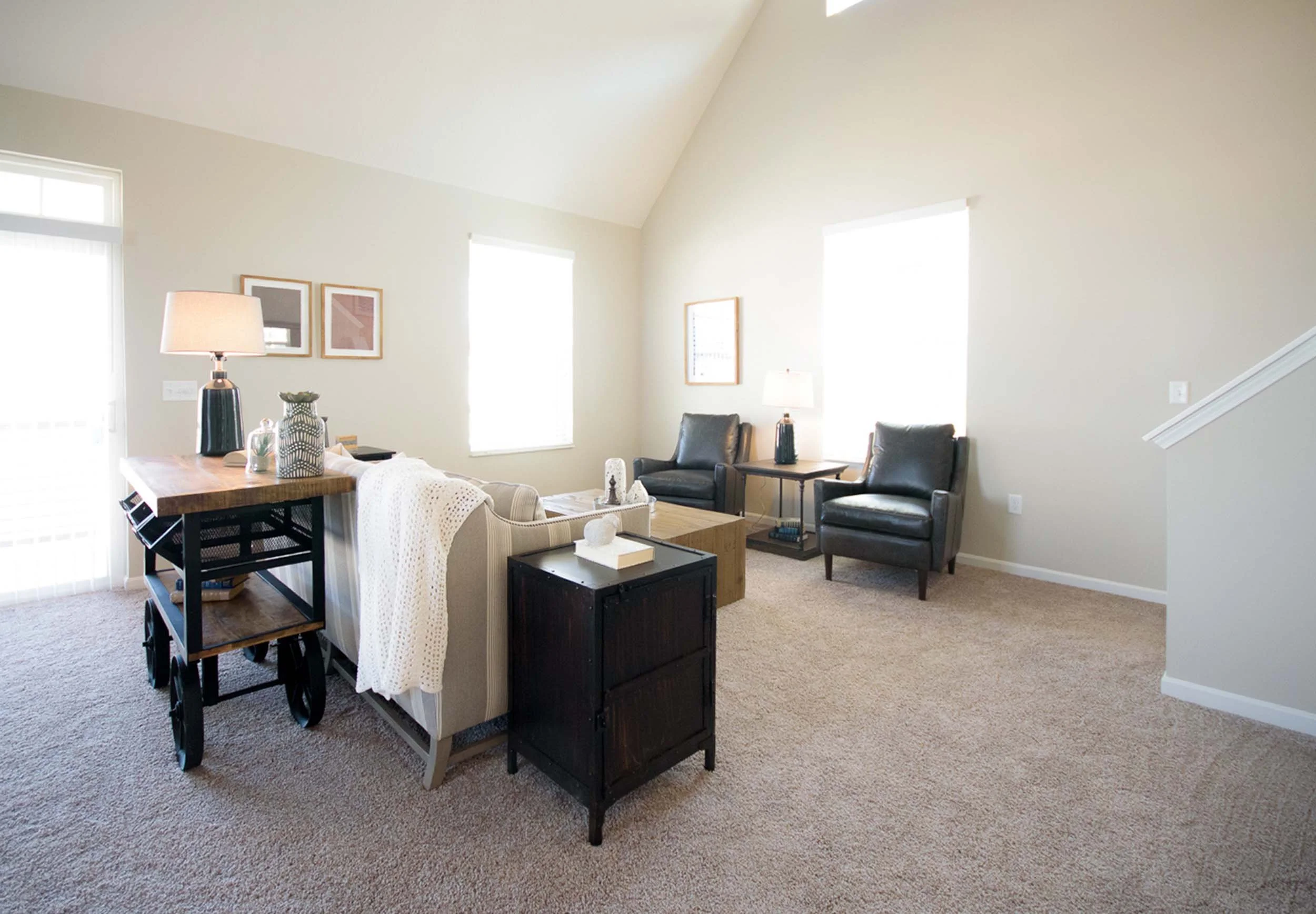 A spacious living room with beige walls, carpeted floor, and two large windows letting in natural light. There are two black armchairs, a small wooden coffee table, a side table with a lamp, and framed art on the walls.