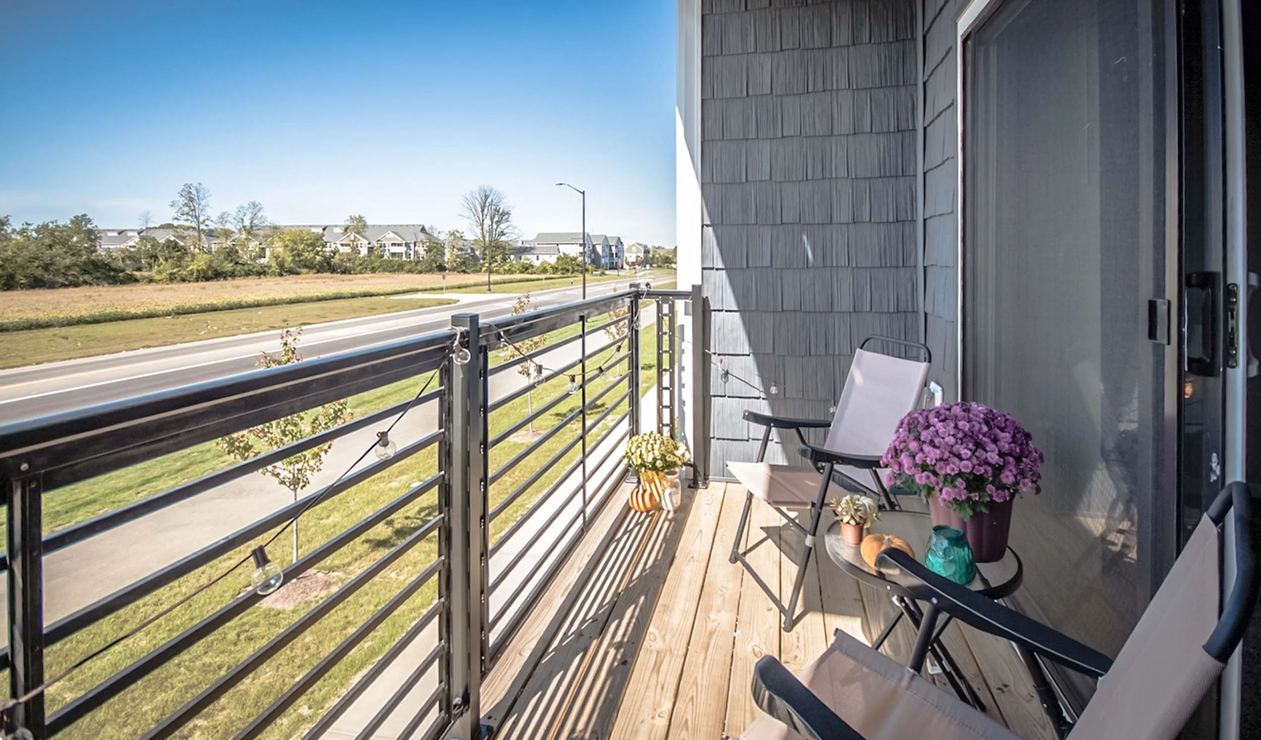 Balcony with flower pots, chairs, and string lights overlooking a street and green lawn under a clear blue sky.