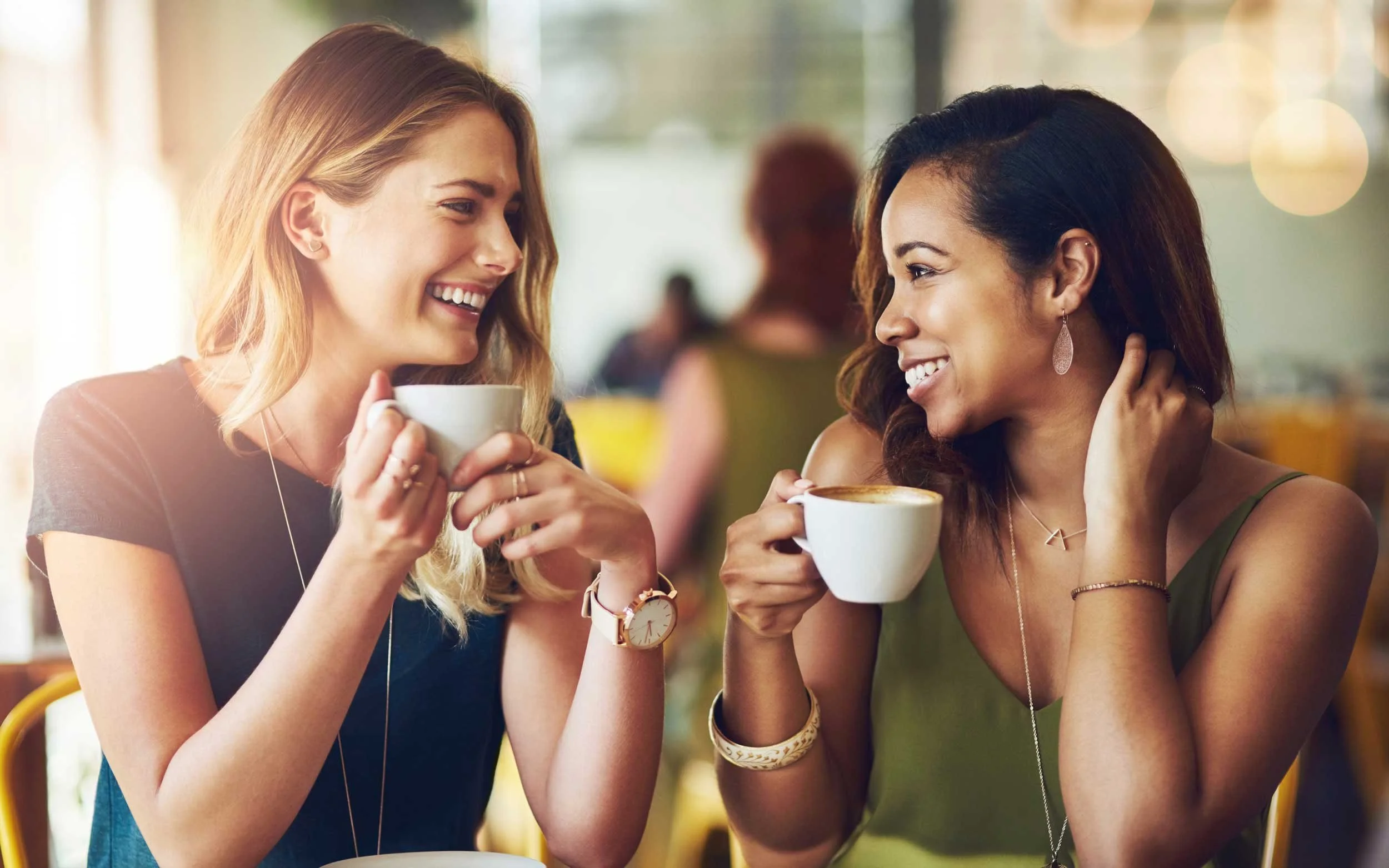 Two women sit at a table in a cafe, smiling and enjoying coffee together. They are looking at each other warmly. The background is blurred with warm lighting.