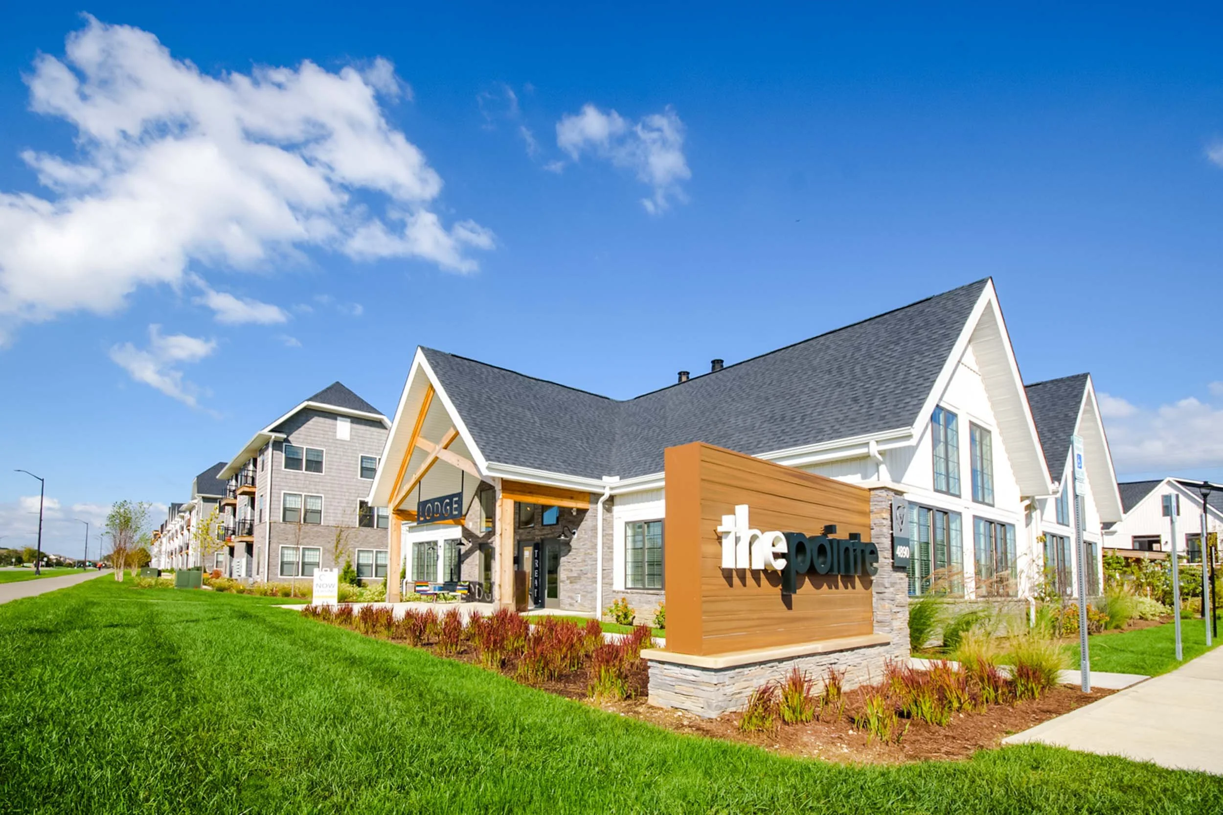 Modern multi-story buildings with a sign reading "the pointe" in a landscaped area with green grass and a blue sky with white clouds.