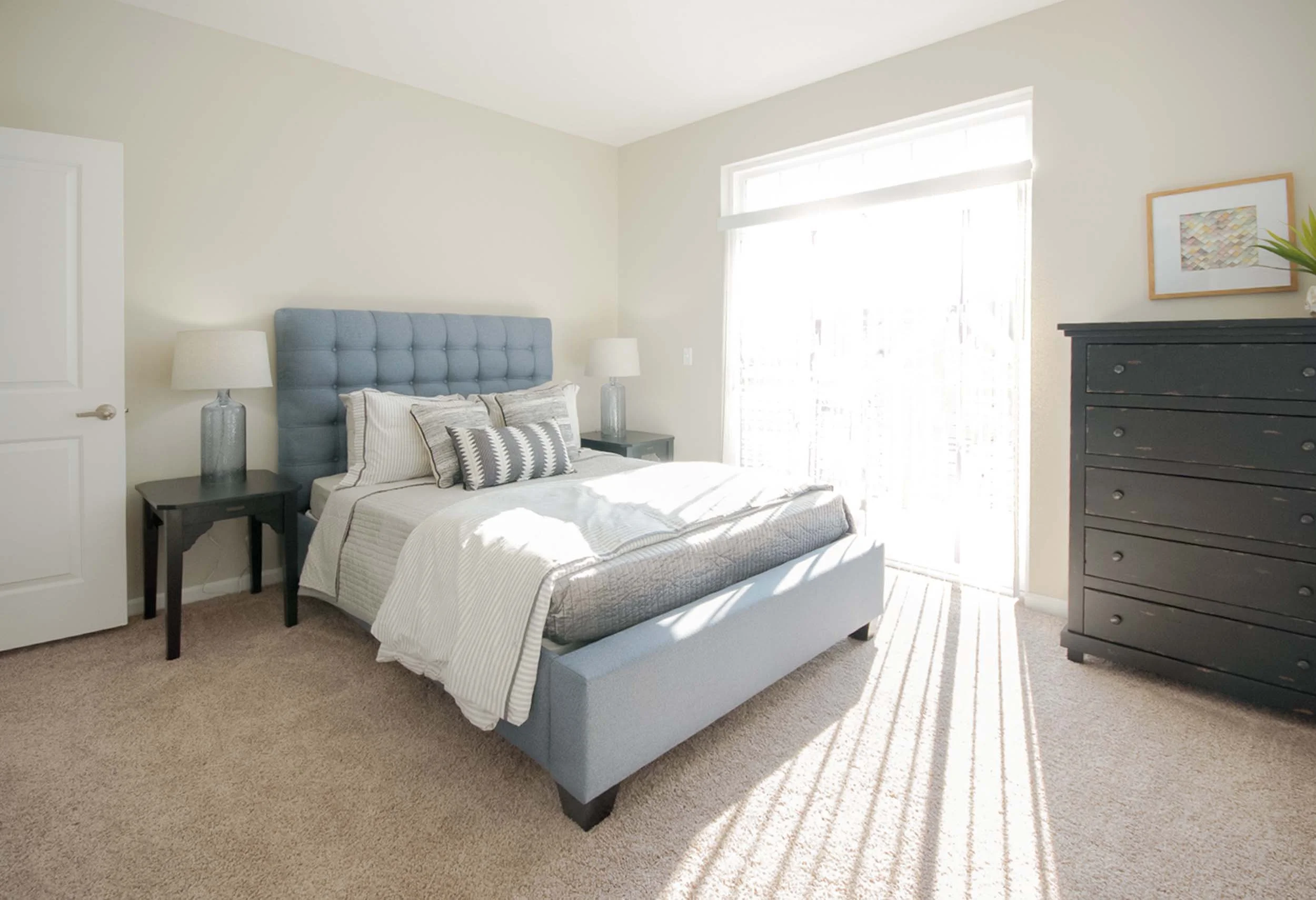 A neatly made bed with a light blue upholstered headboard, striped and patterned pillows, flanked by black bedside tables with lamps, in a bright bedroom with a large window, a black dresser, and a framed artwork on the wall.