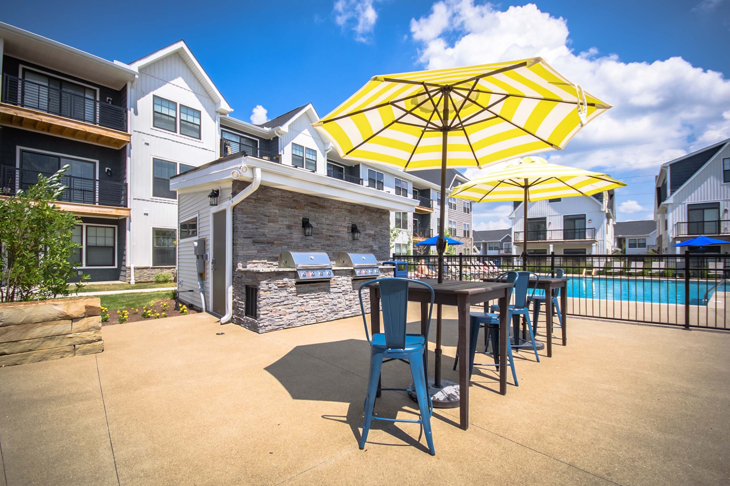 Outdoor pool area with yellow and white striped umbrellas, blue chairs, BBQ grills, and modern apartment buildings in the background under a bright blue sky.