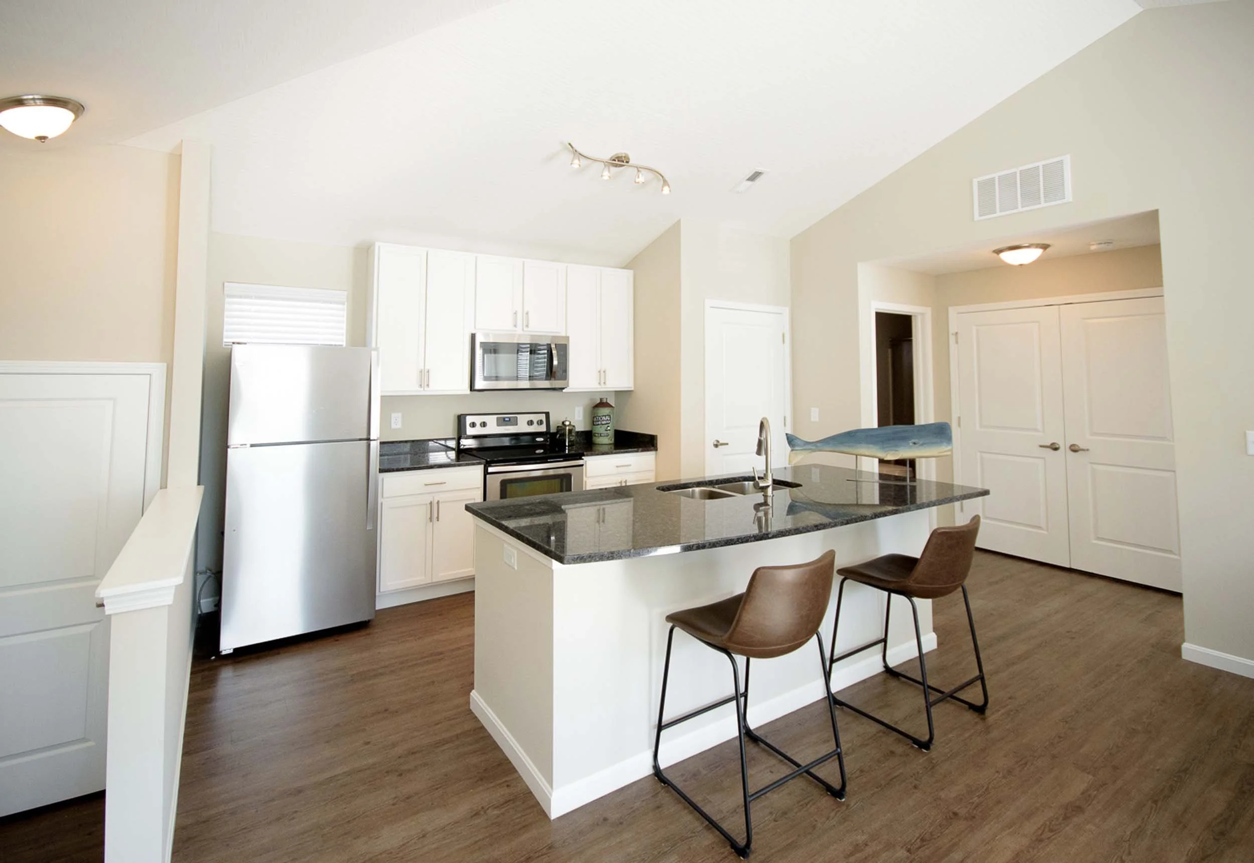 Open-concept kitchen with white cabinets, stainless steel appliances, dark granite countertops, and a breakfast bar with two brown chairs. Wooden flooring and neutral wall colors.