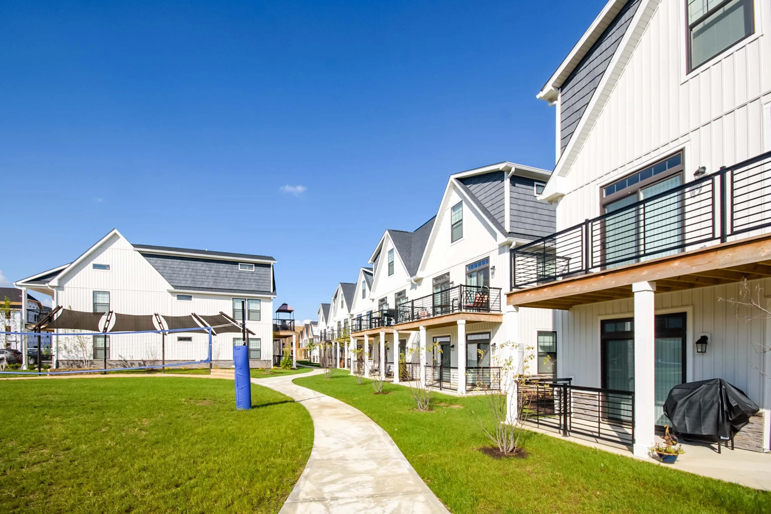 A row of modern white apartment buildings with balconies, set beside a well-maintained green lawn with a winding concrete pathway, under a bright blue sky.