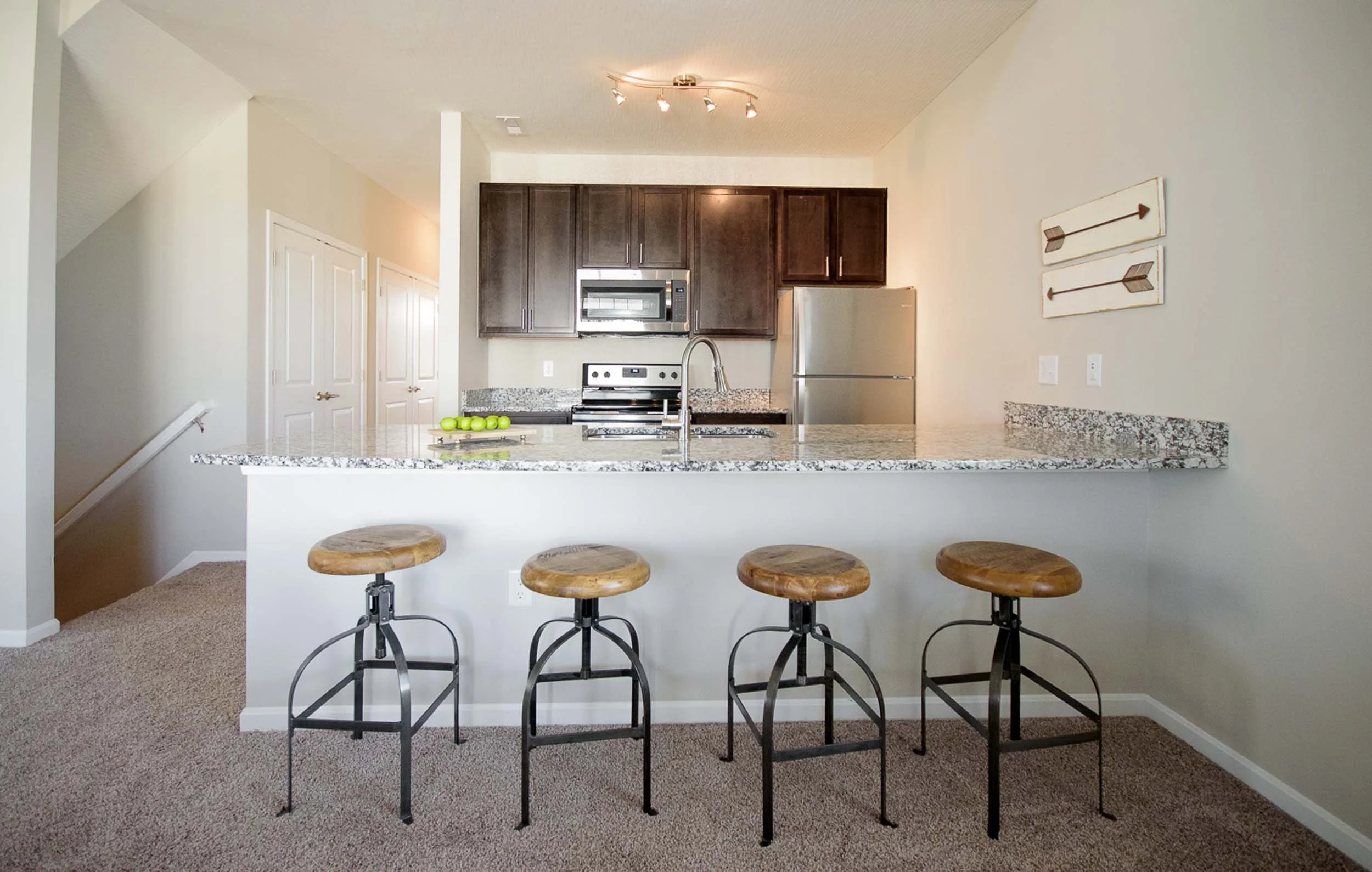 Cozy kitchen with granite countertops, dark wood cabinets, stainless steel appliances, and a breakfast bar with four wooden stools. Decor includes two arrow wall art pieces.