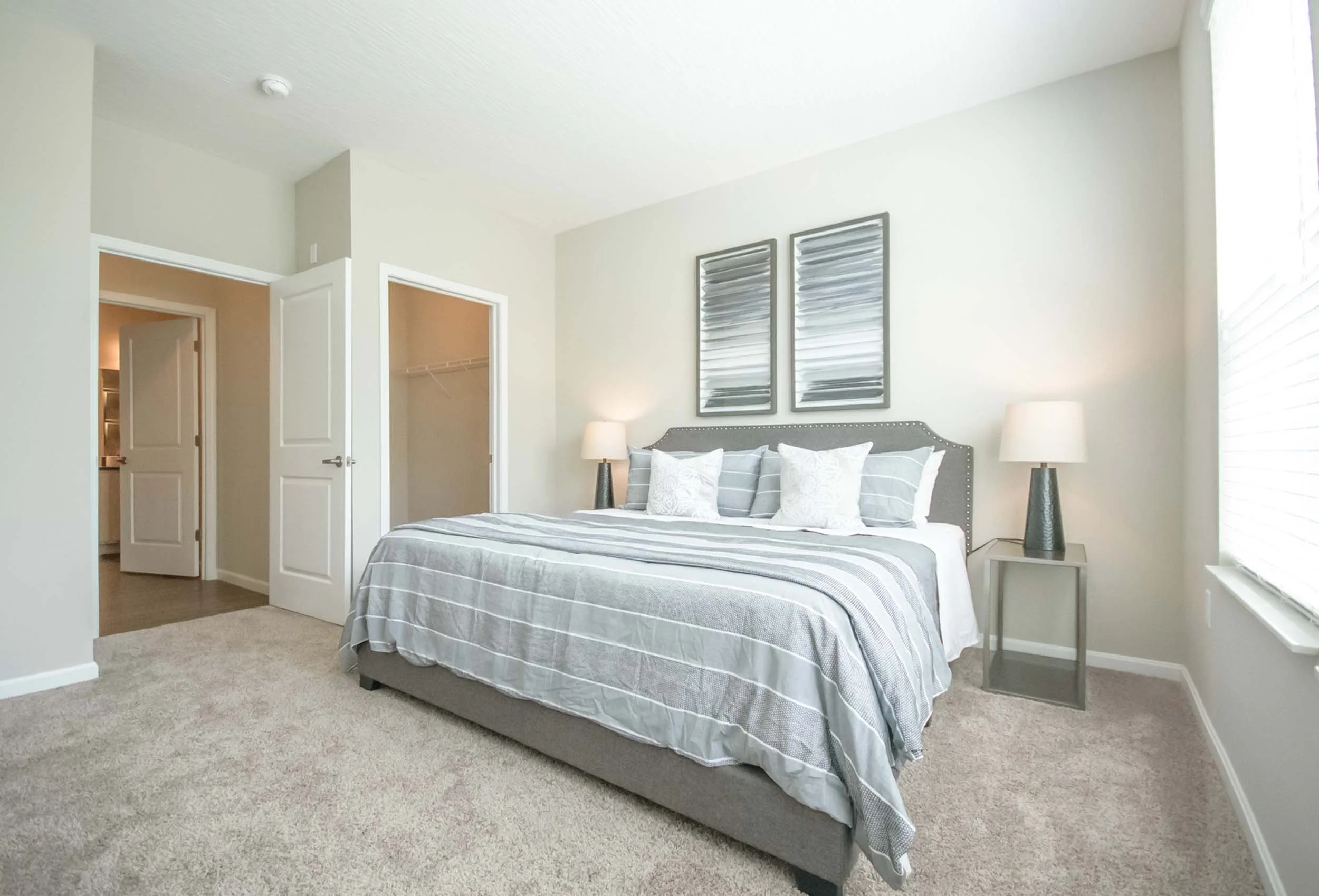 A bedroom with a bed, two nightstands with lamps, framed artwork above the bed, a closet with open doors, and a window with blinds.