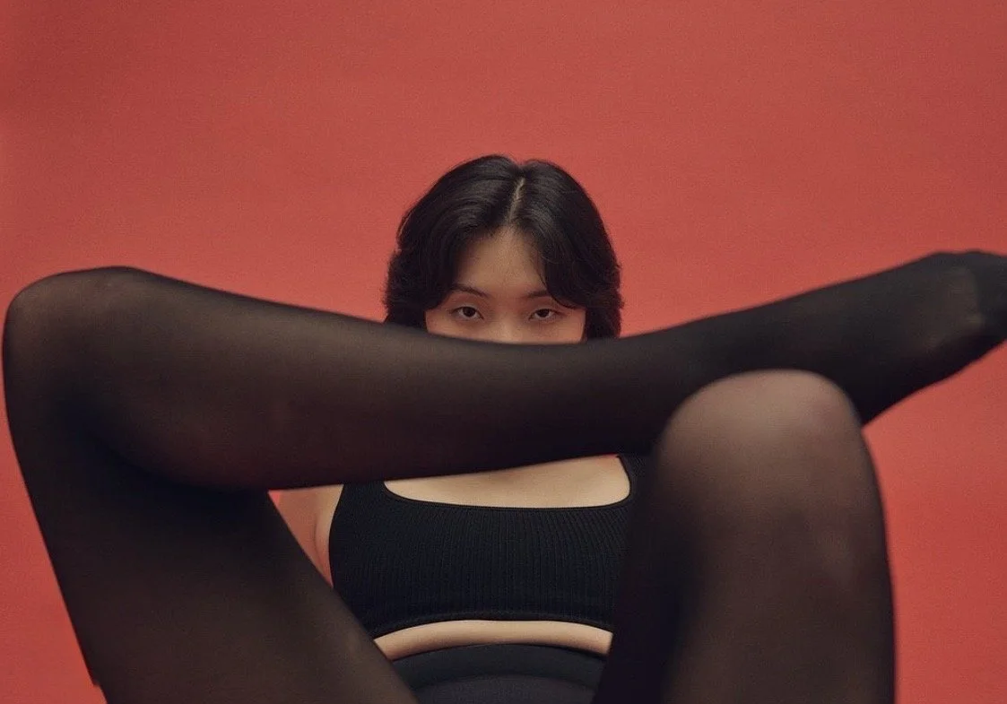 A woman with short black hair, wearing a black sports bra and black tights, sitting against a red background, with her legs spread and her arms extended, partially covering her face.