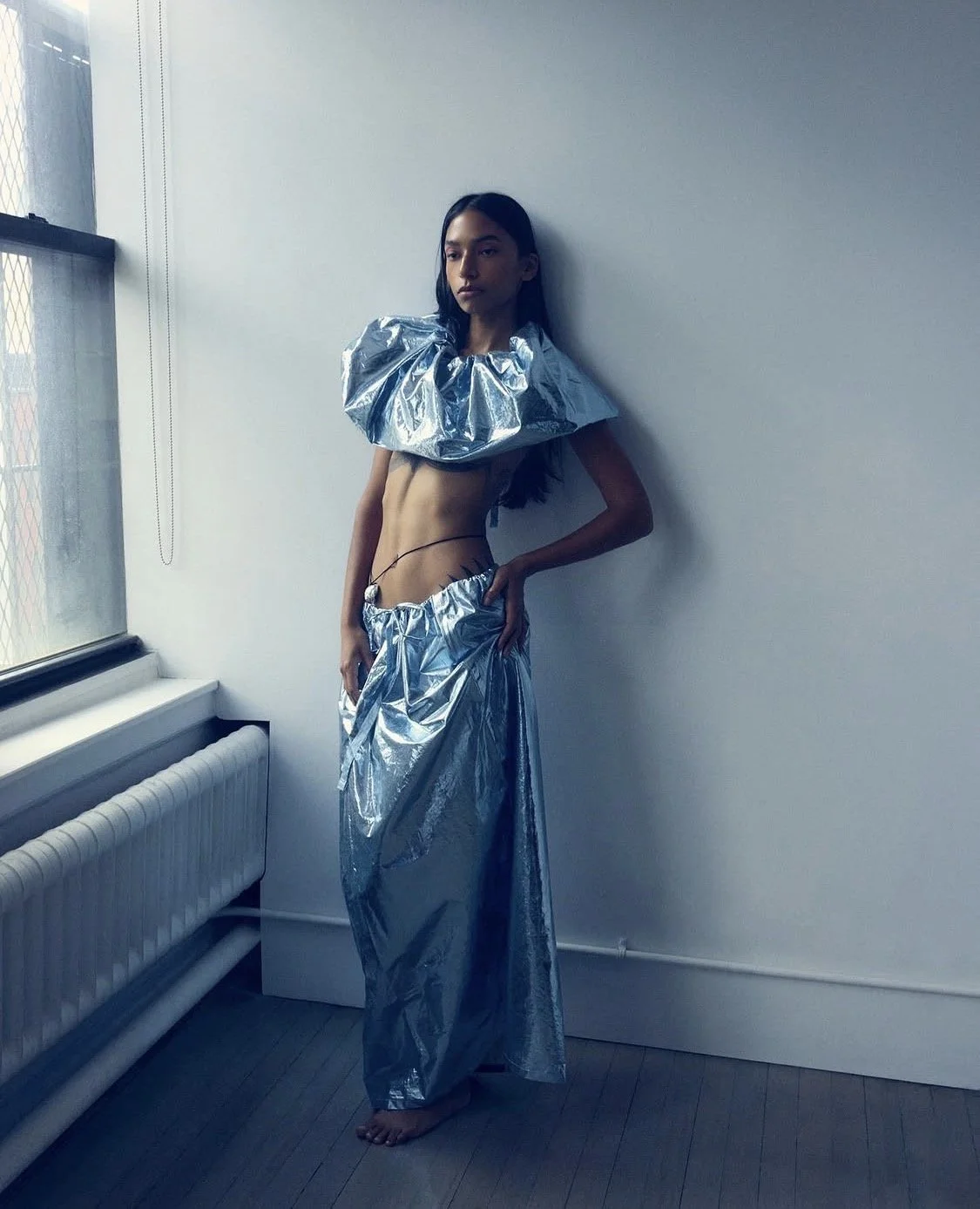 A woman with long dark hair standing barefoot against a white wall, wearing a futuristic metallic silver outfit with large ruffled shoulder pieces and a long skirt, near a window with a metal grid.