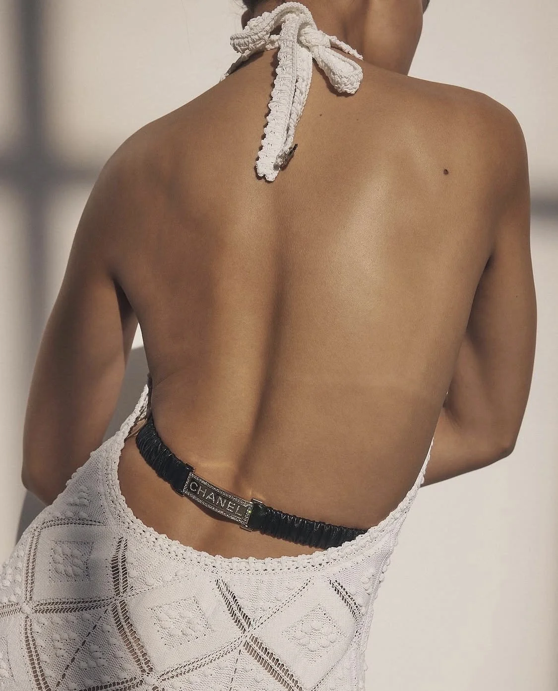 Close-up of a person's back showing a white knitted halter top secured with a knot, with a black belt featuring a Chanel logo around the waist.