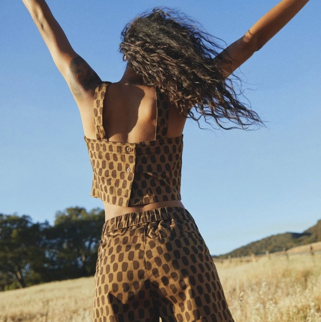 A woman with curly hair standing in an open field with her arms raised, wearing a brown and black patterned sleeveless top and matching pants, under a clear blue sky.