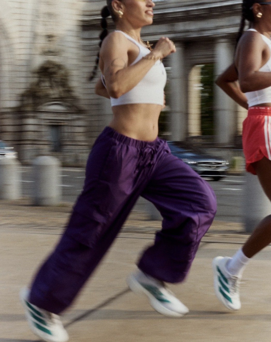 A woman running outdoors wearing purple track pants, a white crop top, white sneakers, and gold earrings, with another runner partially visible next to her.
