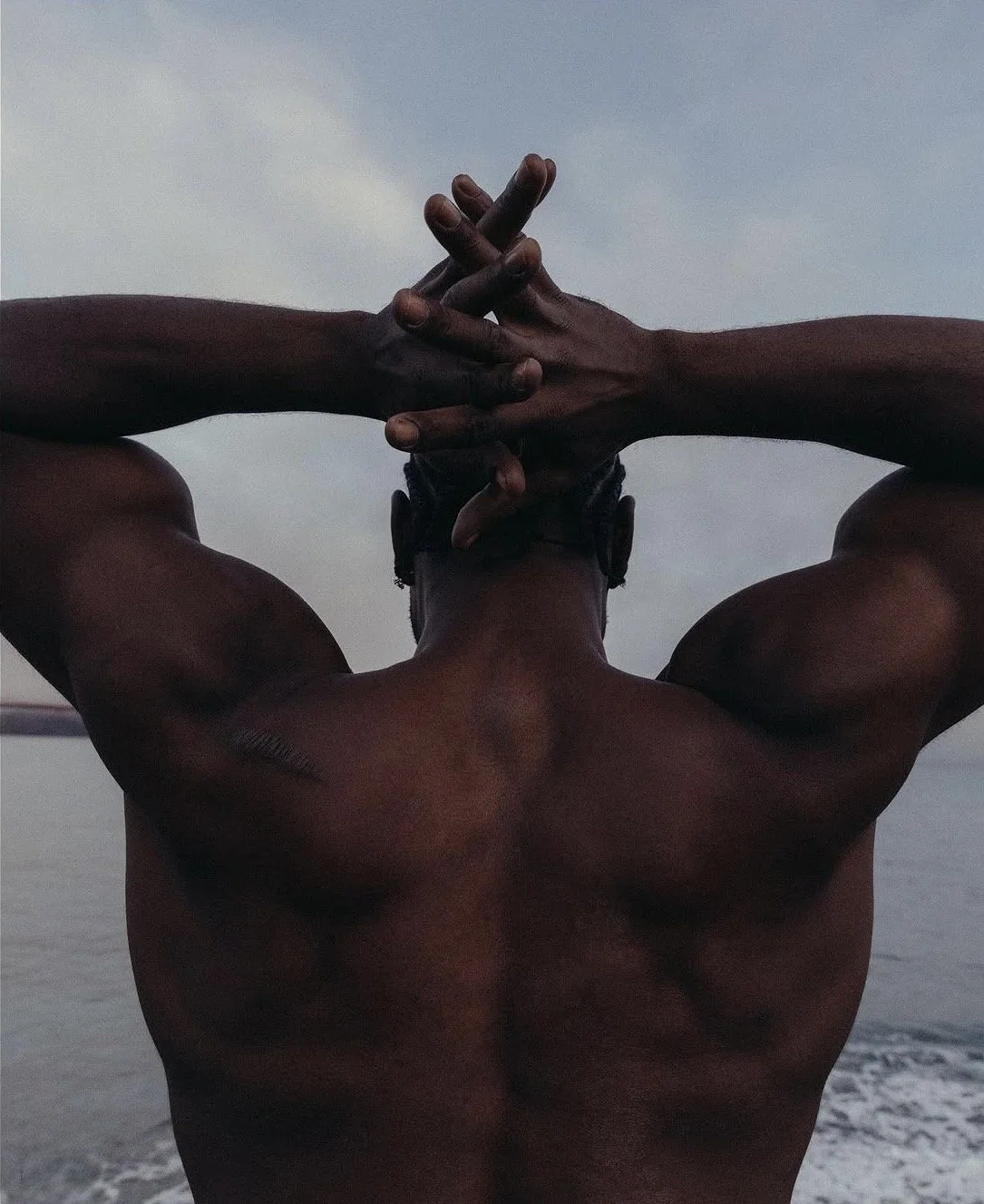 Back view of a shirtless man with muscular arms, standing outdoors by the water, with hands clasped behind his head and head slightly tilted back.