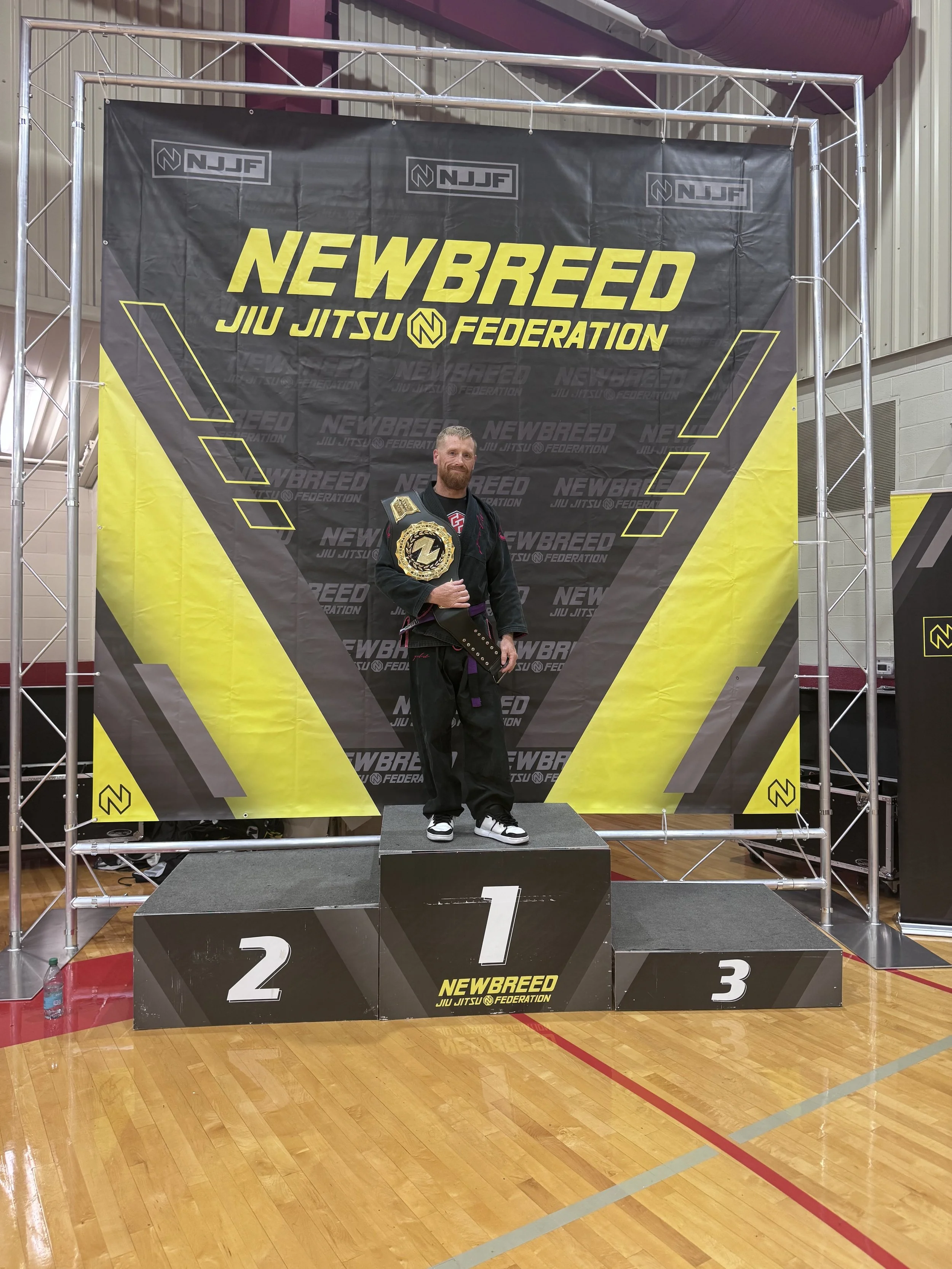 A man standing on a winners' podium with a championship belt, at a martial arts competition, under a black and yellow 'Newbreed Jiu Jitsu Federation' banner.
