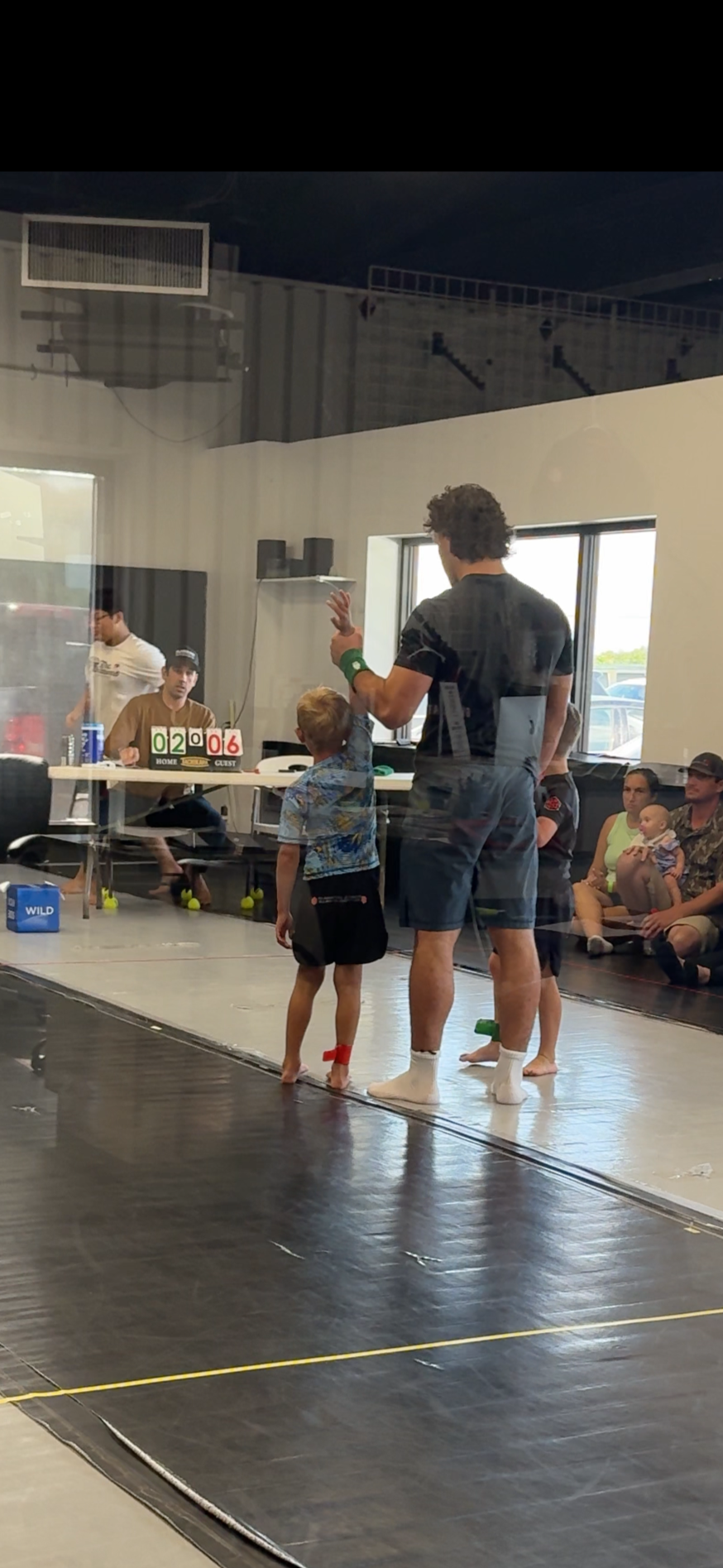 A man and a boy talking inside a room at a sporting event, with several seated spectators observing, a scoreboard indicating 2-0-6, and tennis balls on the floor.