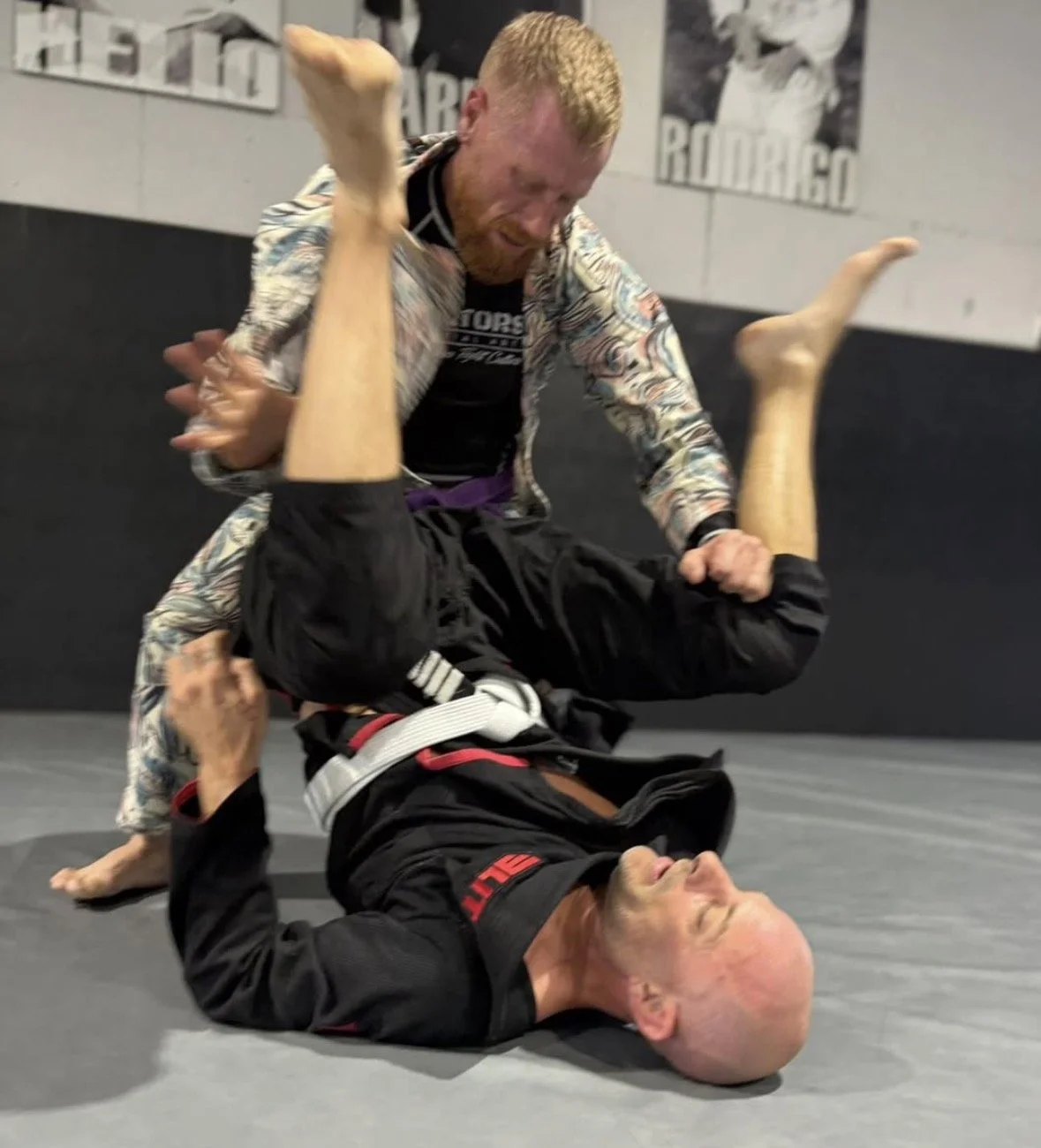 Two men practicing Brazilian Jiu-Jitsu on a mat. One is in a black gi on the ground, holding the other in a chokehold, while the other man is in a colorful rash guard, attempting to escape.