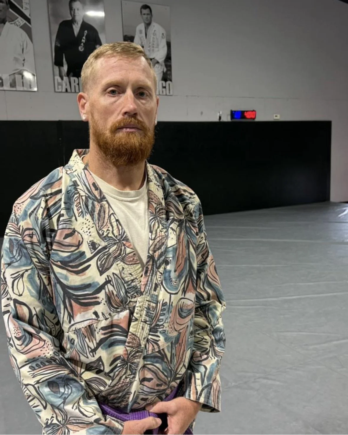 A man with red hair and beard wearing a colorful, patterned long sleeve shirt standing in a martial arts training area.