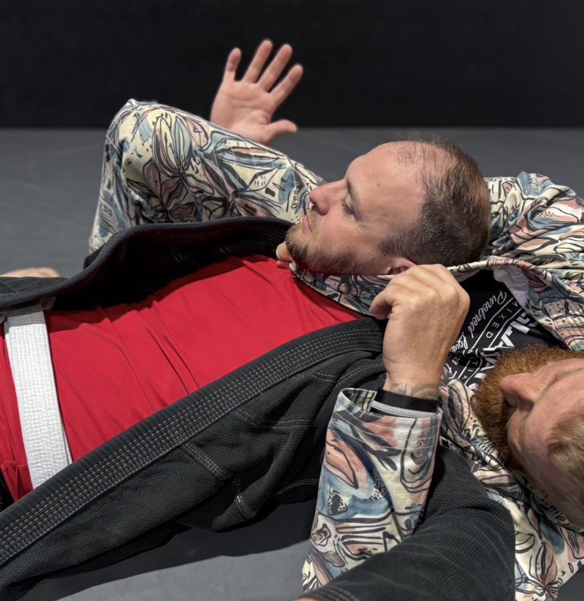 Two men practicing Brazilian Jiu-Jitsu on a mat, one lying on his back with a black gi and red shirt, the other partially visible wearing a colorful gi, both focusing on their grappling positions.