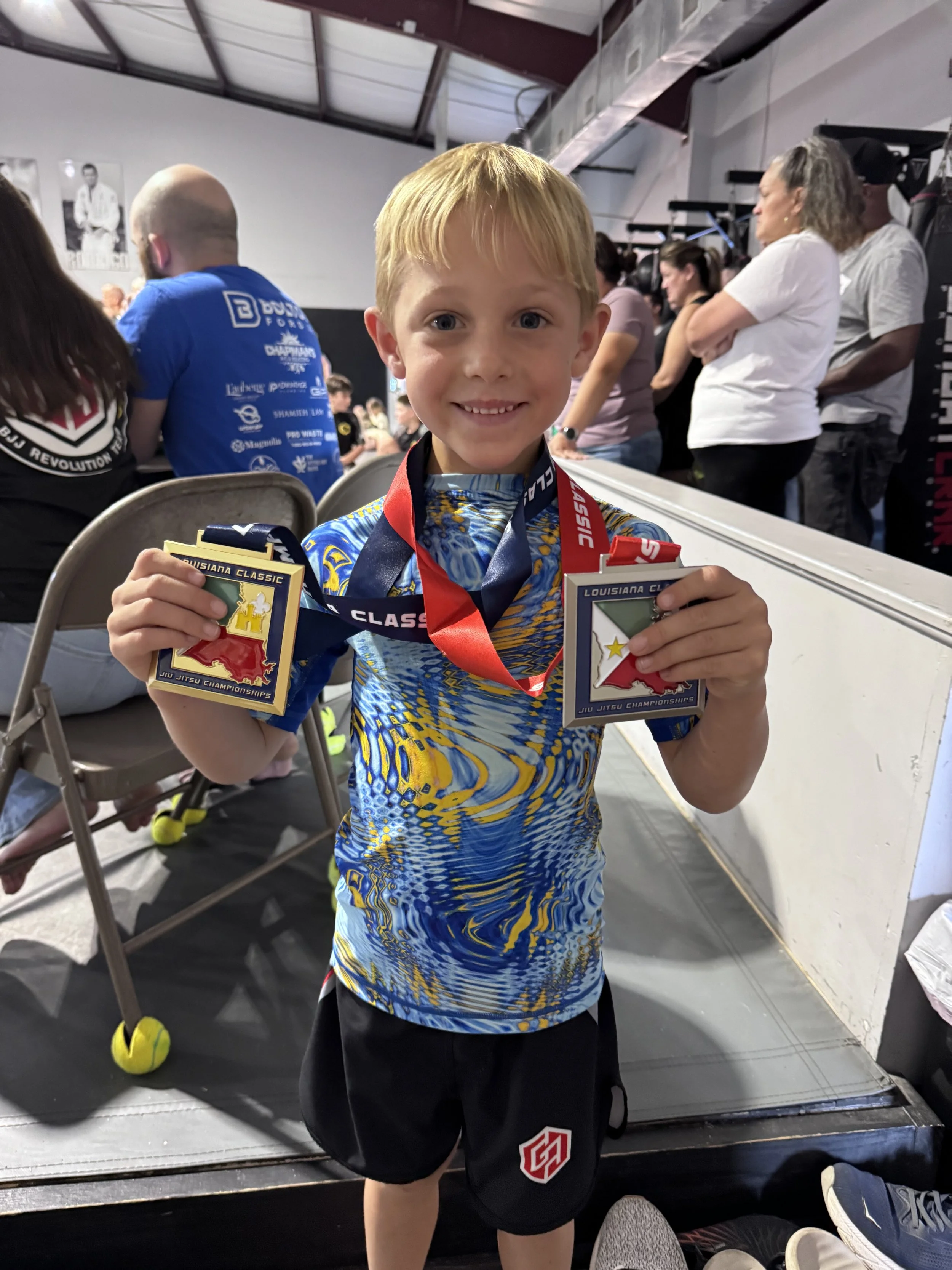 A young boy with blond hair and a bright smile is holding two medals in his hands, standing in front of a boxing ring or martial arts competition area. He is wearing athletic clothing with a blue, yellow, and white pattern and black shorts with a red and white shield logo. The background shows several people, some sitting and some standing, in a gym or sports arena setting.