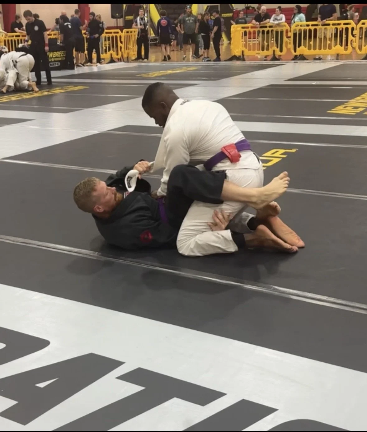 Two men practicing Brazilian Jiu-Jitsu on a mat at a martial arts competition. One man in a white gi is on top, and the other man in a black gi is on the bottom. In the background, people are watching and preparing for matches.