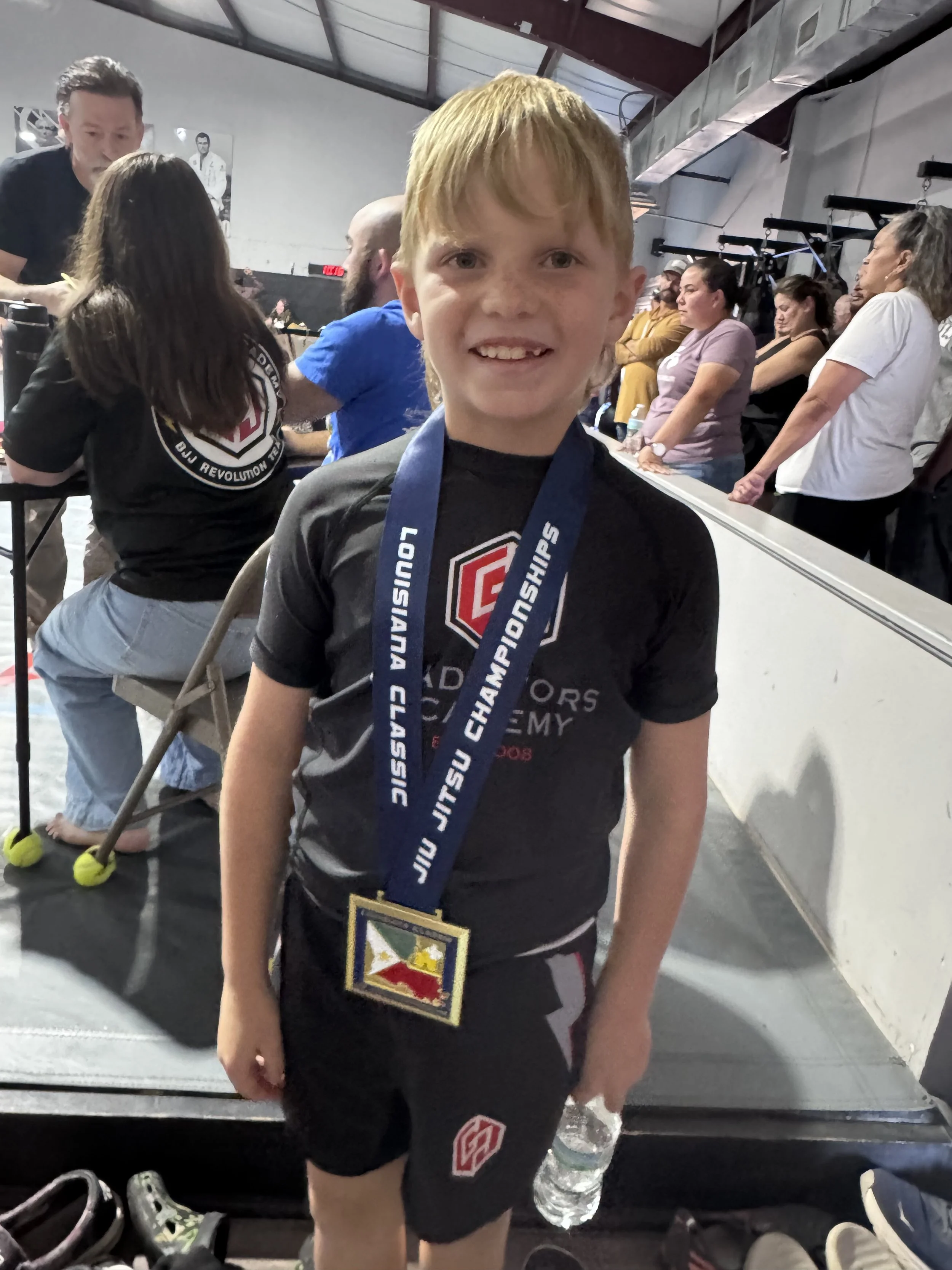 Young boy with a medal around his neck, smiling at the camera, in a gym or training facility with spectators and other individuals in the background.
