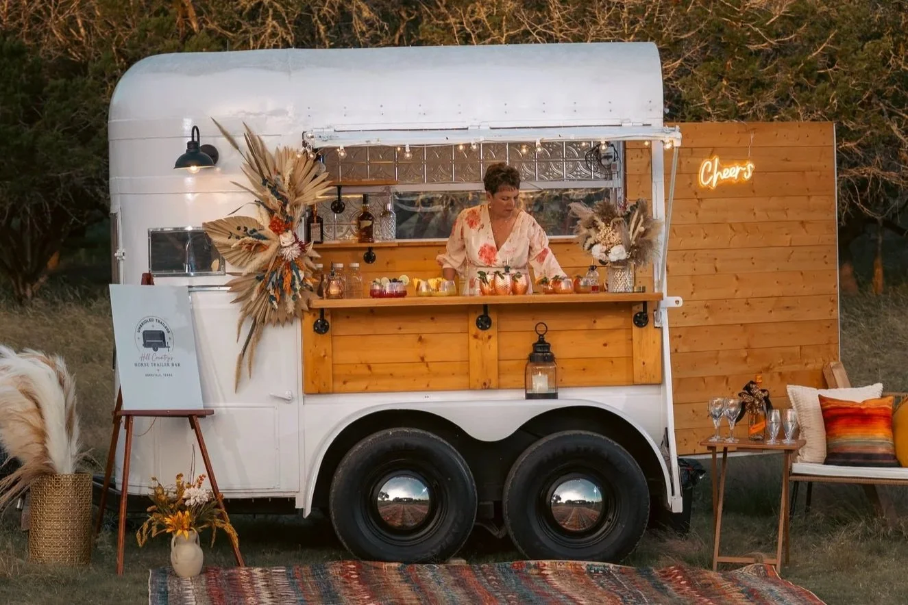 A food truck with a wood counter serving drinks, decorated with dried flowers, situated outdoors with trees in the background. A woman stands behind the counter preparing beverages. To the right, a neon sign says 'Cheers,' and there's a small seating
